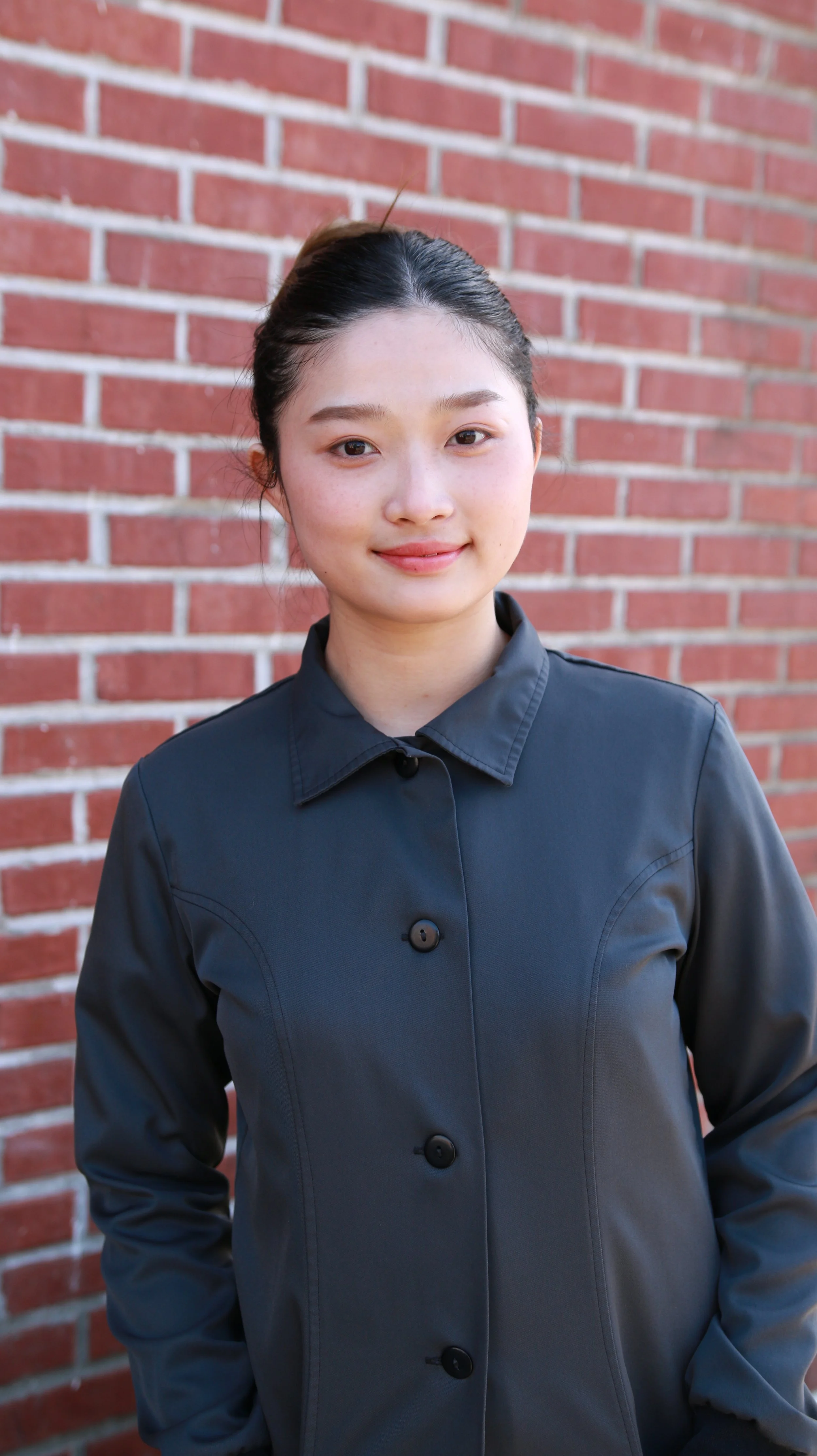 Young woman with dark hair in a bun, wearing a dark button-up shirt, standing in front of a brick wall.
