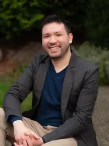 A man sitting outdoors, smiling, wearing a dark blazer and a navy blue shirt, with greenery in the background.