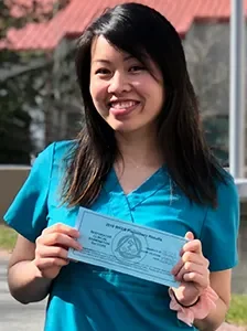 A woman in blue scrubs holding a certificate outdoors, smiling.