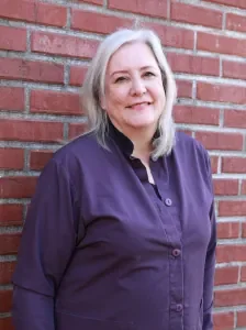 A woman with blonde hair standing in front of a brick wall, wearing a purple shirt and smiling.