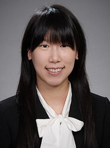 A young woman with long black hair and bangs, wearing a black blazer and white blouse with a bow, smiling at the camera.