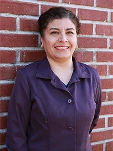 Woman with dark hair smiling, wearing a purple jacket, standing against a brick wall.