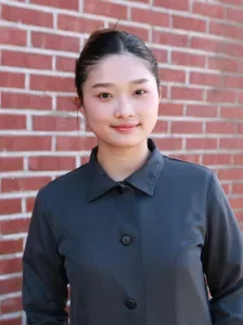 Young woman with dark hair in a bun, wearing a dark button-up shirt, standing in front of a brick wall.