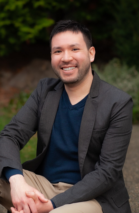 A man sitting outdoors, smiling, wearing a dark blazer and a navy blue shirt, with greenery in the background.
