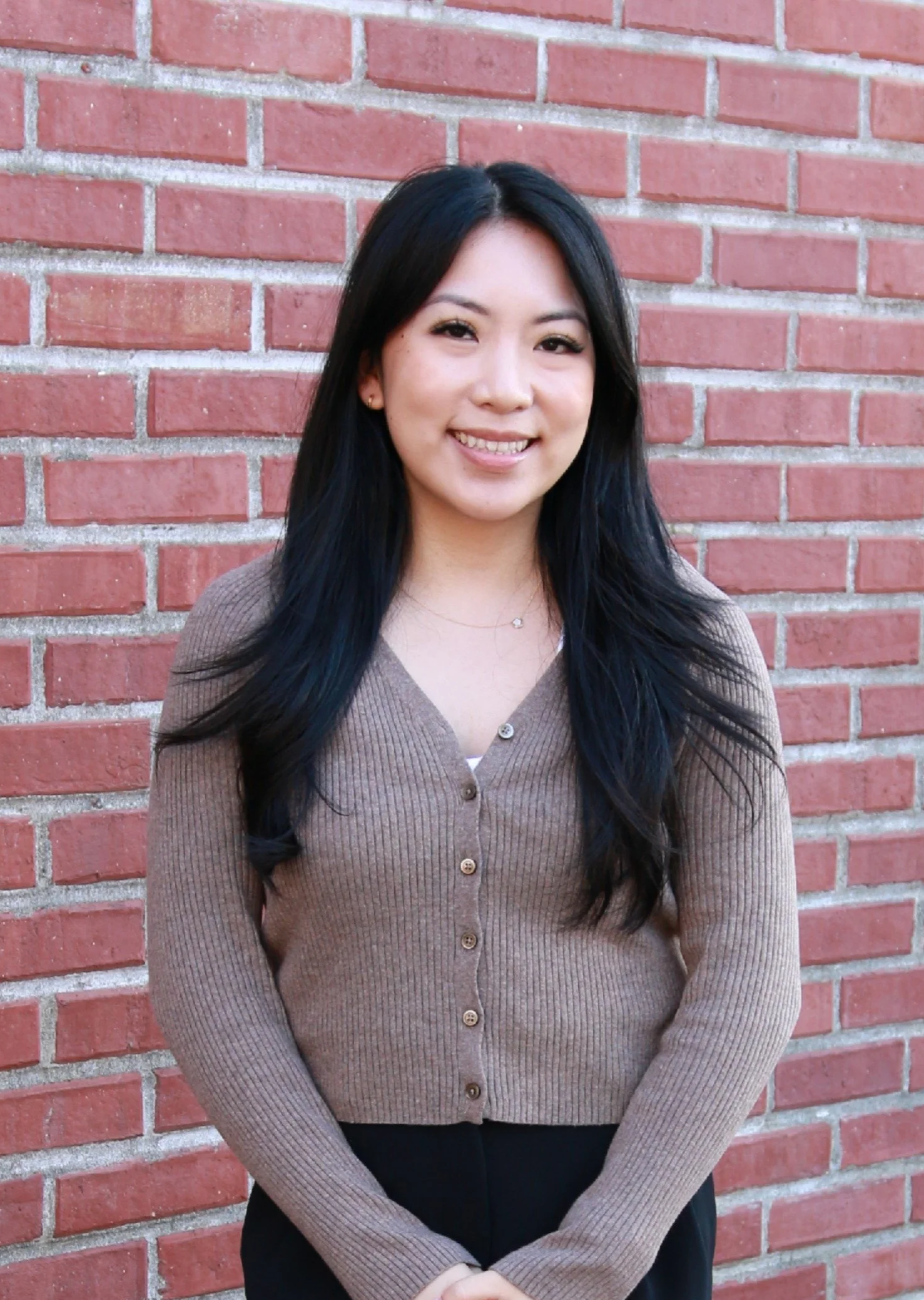 A young woman with long black hair standing in front of a red brick wall, smiling and wearing a beige cardigan.
