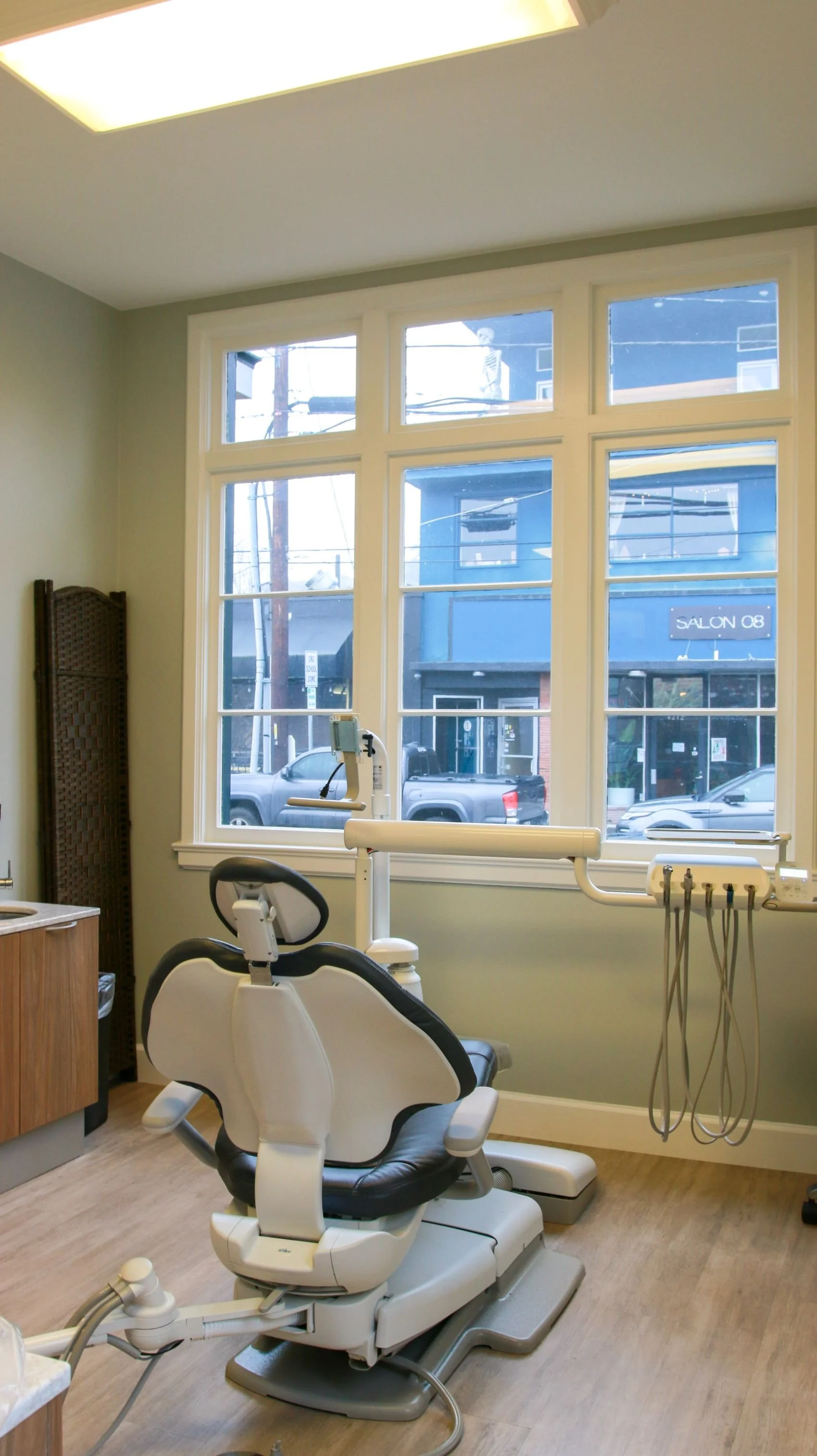 Empty dental office chair in front of large window with street view outside, dental equipment nearby.