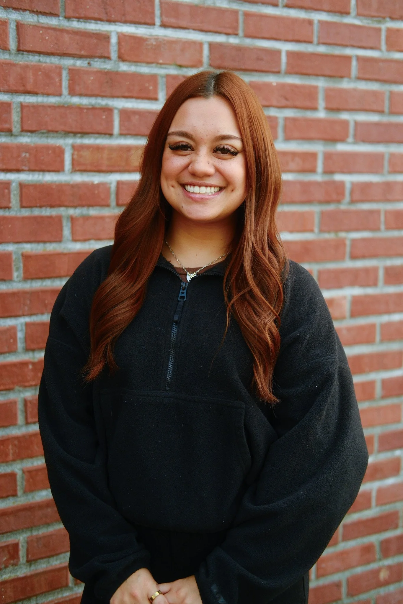 A young woman with long dark hair smiling, standing in front of a brick wall, wearing a black top and a pink striped shirt.