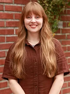 Portrait of a young woman with long red hair smiling, wearing a brown button-up shirt, standing in front of a brick wall with greenery.