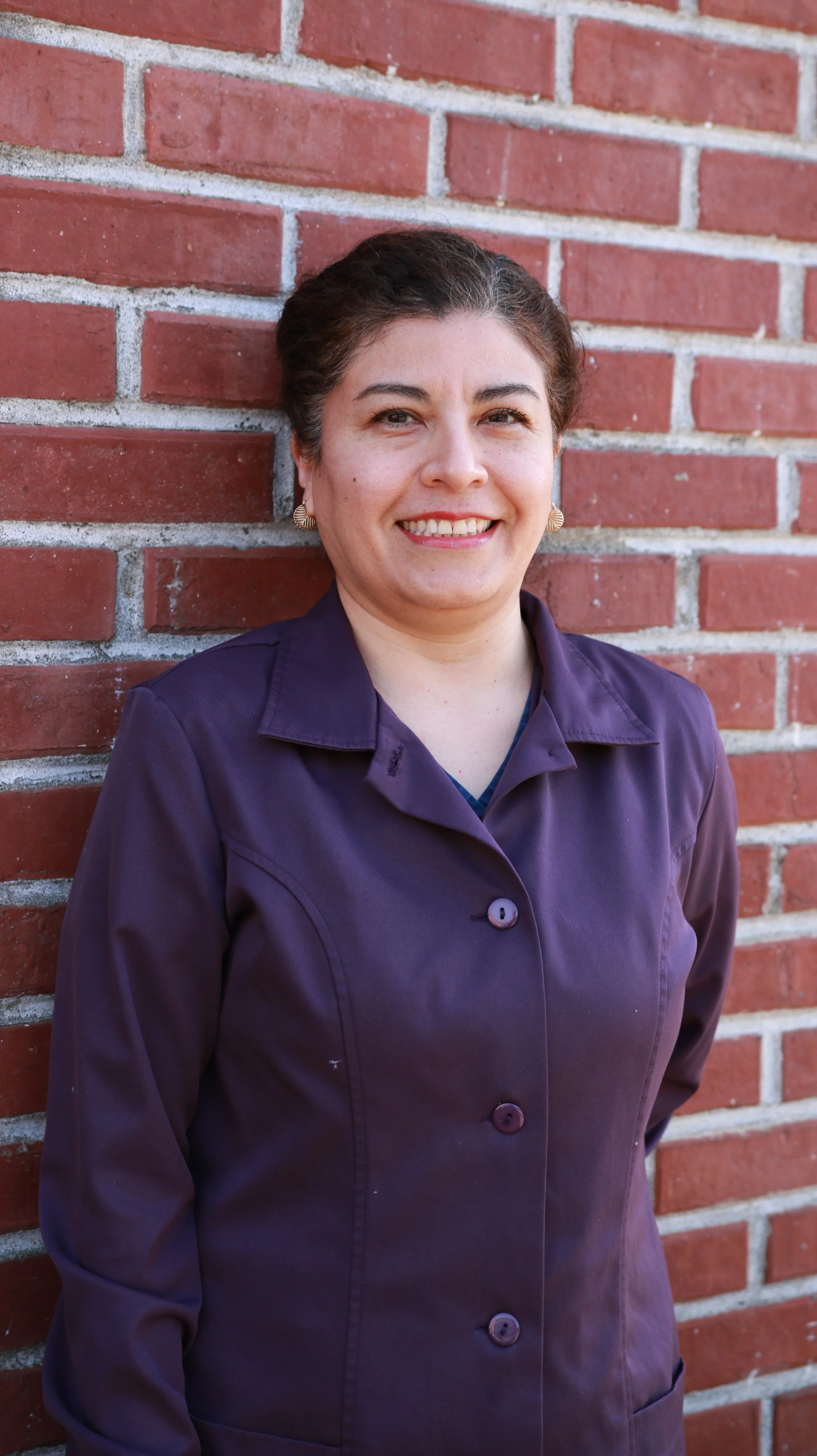 Woman with dark hair smiling, wearing a purple jacket, standing against a brick wall.