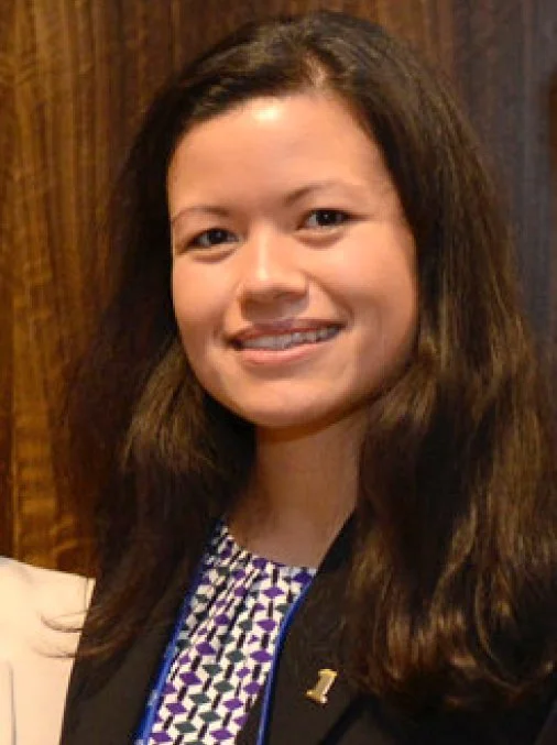 A smiling young woman with long dark hair, wearing a patterned blouse and black blazer, standing in front of a wooden background.