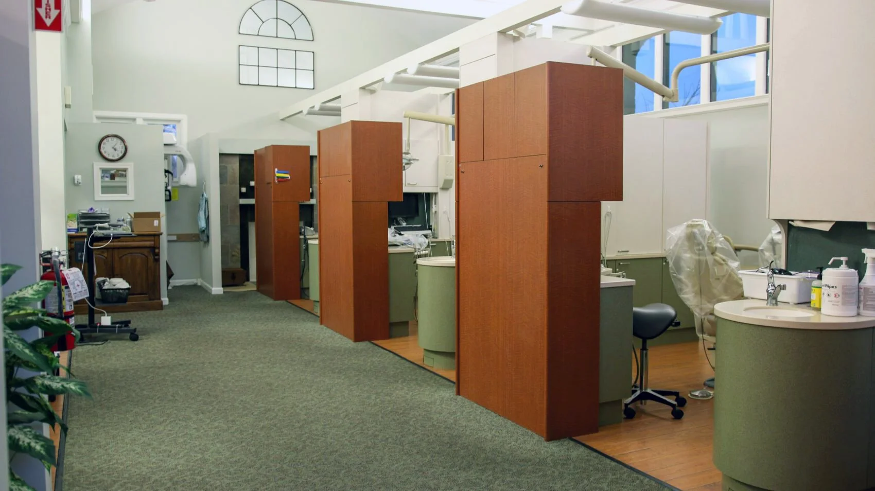 Hospital reception area with multiple private patient examination rooms separated by wooden partitions, medical supplies on a counter, and a sink, with large windows and a clock on the wall.