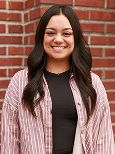 A young woman with long dark hair smiling, standing in front of a brick wall, wearing a black top and a pink striped shirt.