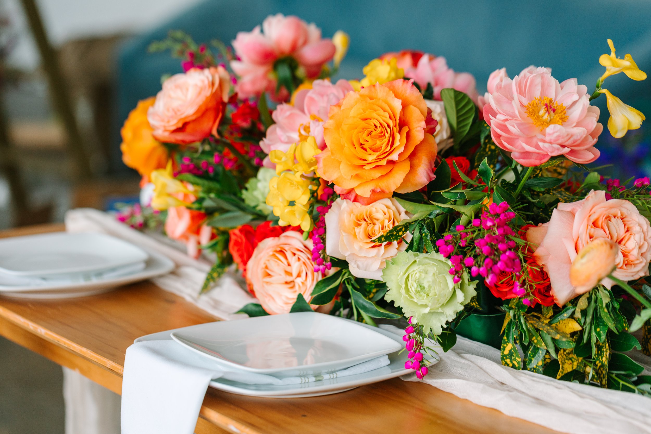 A wooden dining table set with white plates and napkins features a large, colorful floral centerpiece with pink, peach, yellow, and purple flowers and green leaves.