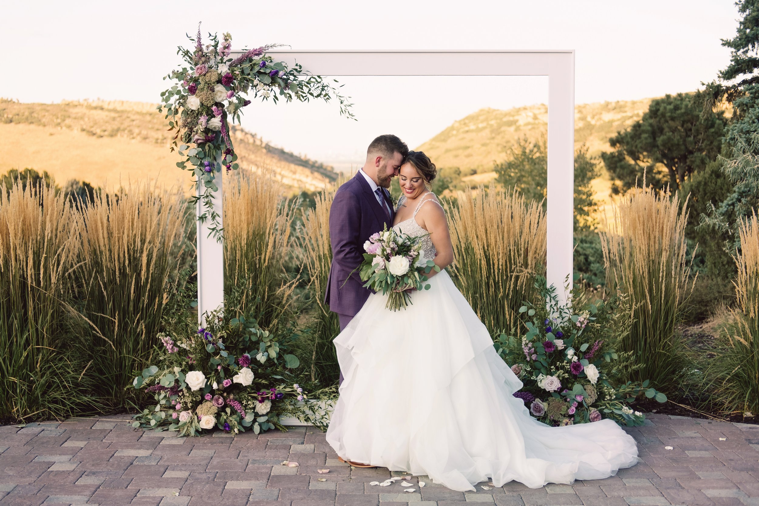 Bride and groom at outdoor wedding, standing close under white floral arch with purple, white, and green flowers, in a natural setting with mountains and tall grasses.