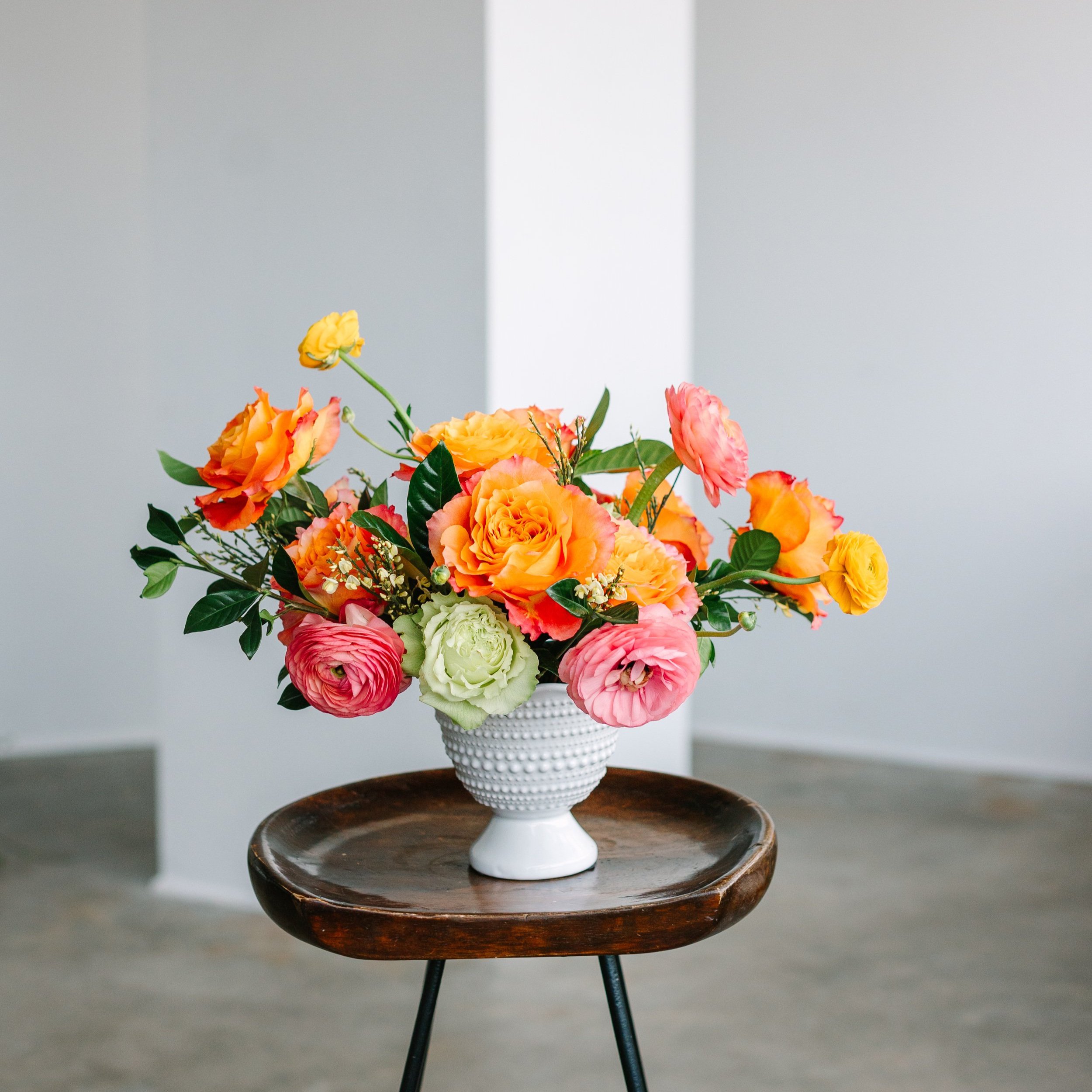 A bouquet of colorful flowers including roses and ranunculus in a white textured vase on a wooden table.