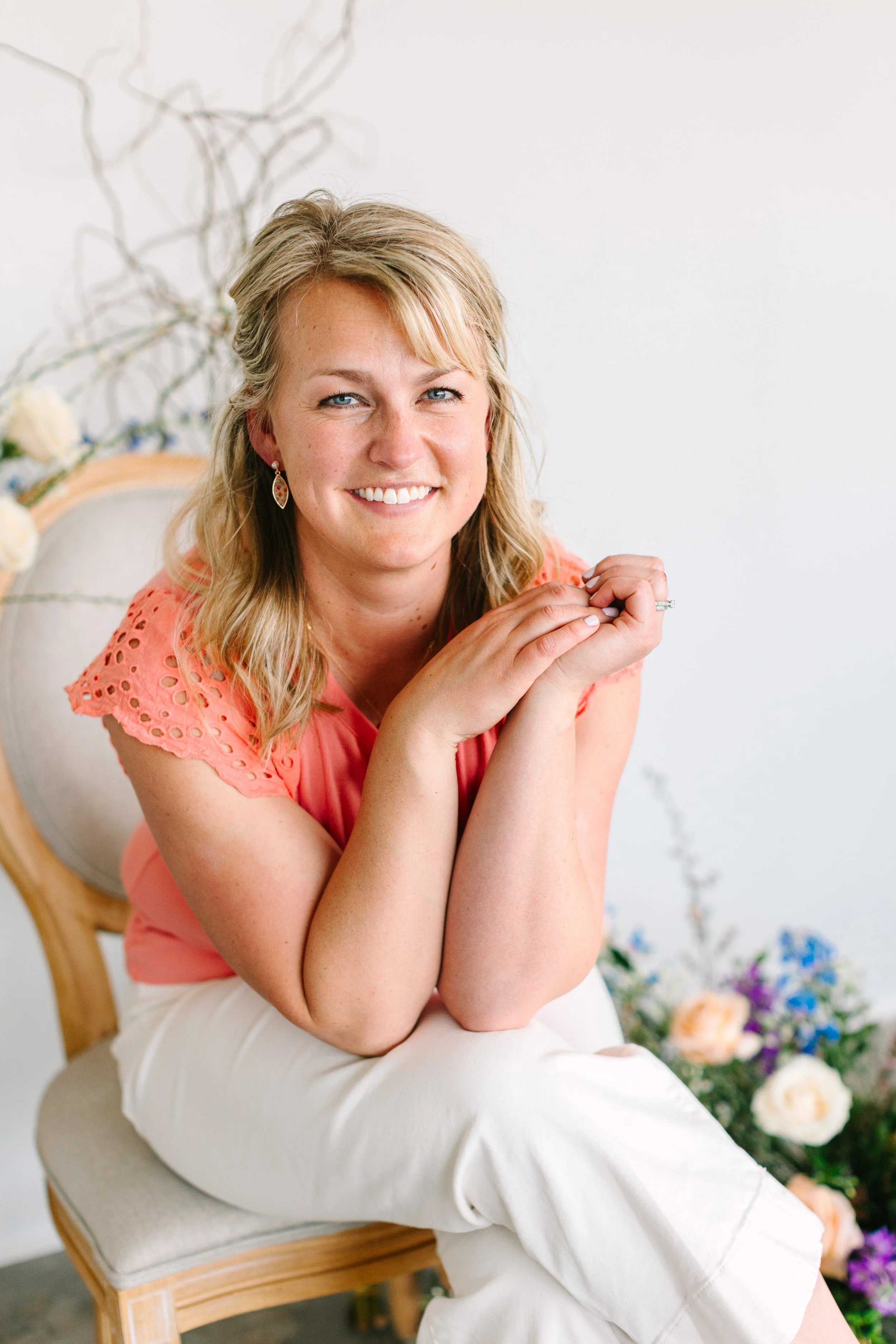 A woman with blonde hair, smiling, wearing a coral blouse with eyelet details on the sleeves, sitting on a wooden chair with a cushioned backrest, in front of a floral arrangement with white, pink, and purple flowers.