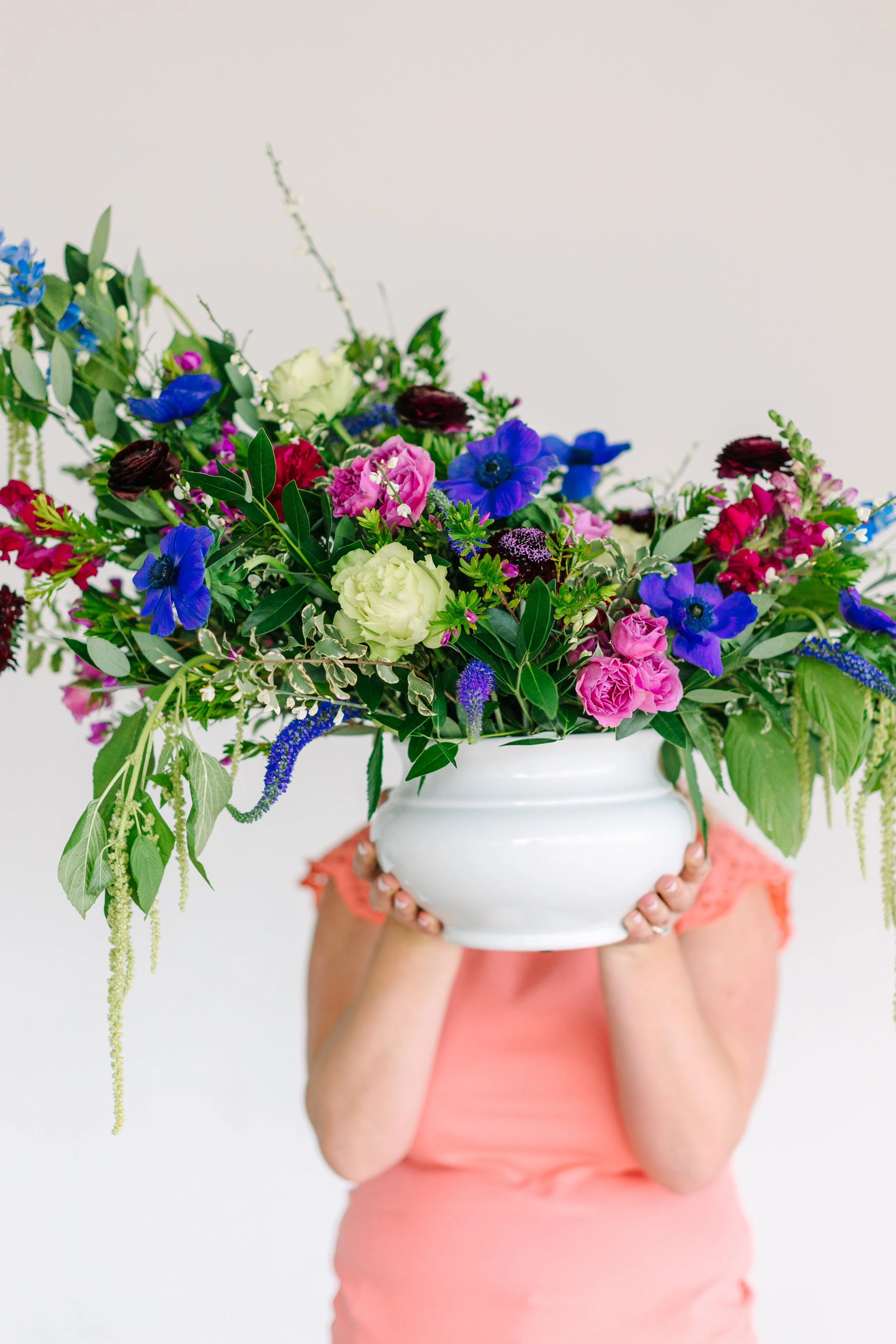 Person holding a large white vase filled with colorful flowers including blue, pink, and cream blooms, with the person wearing a pink top.