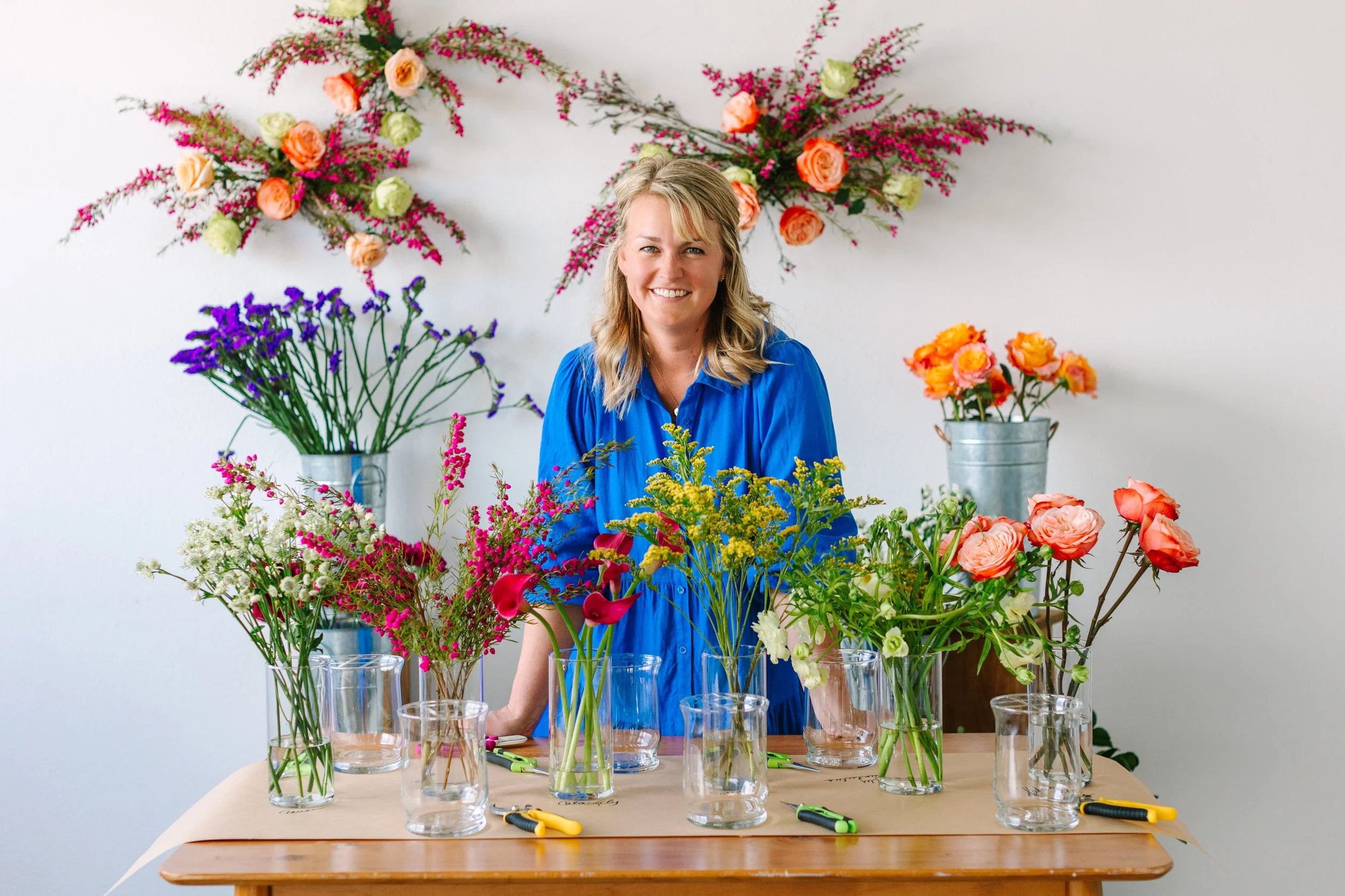 A woman in a blue shirt smiling at a table filled with various colorful flowers in glass vases, with floral arrangements on the wall behind her, including roses and other blossoms.