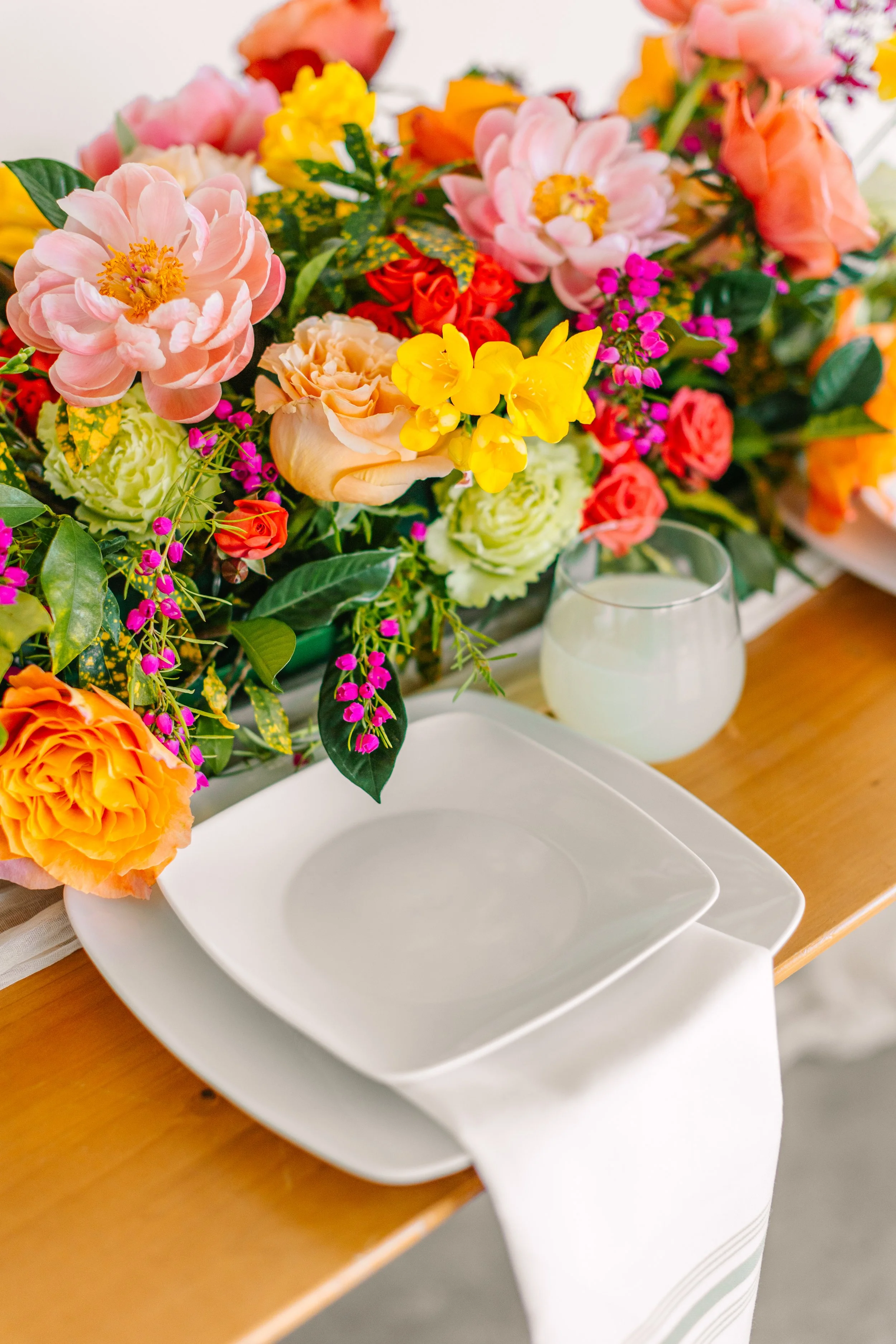 Colorful arrangement of pink, orange, yellow, and purple flowers on a table with white dishware and a glass of white wine.