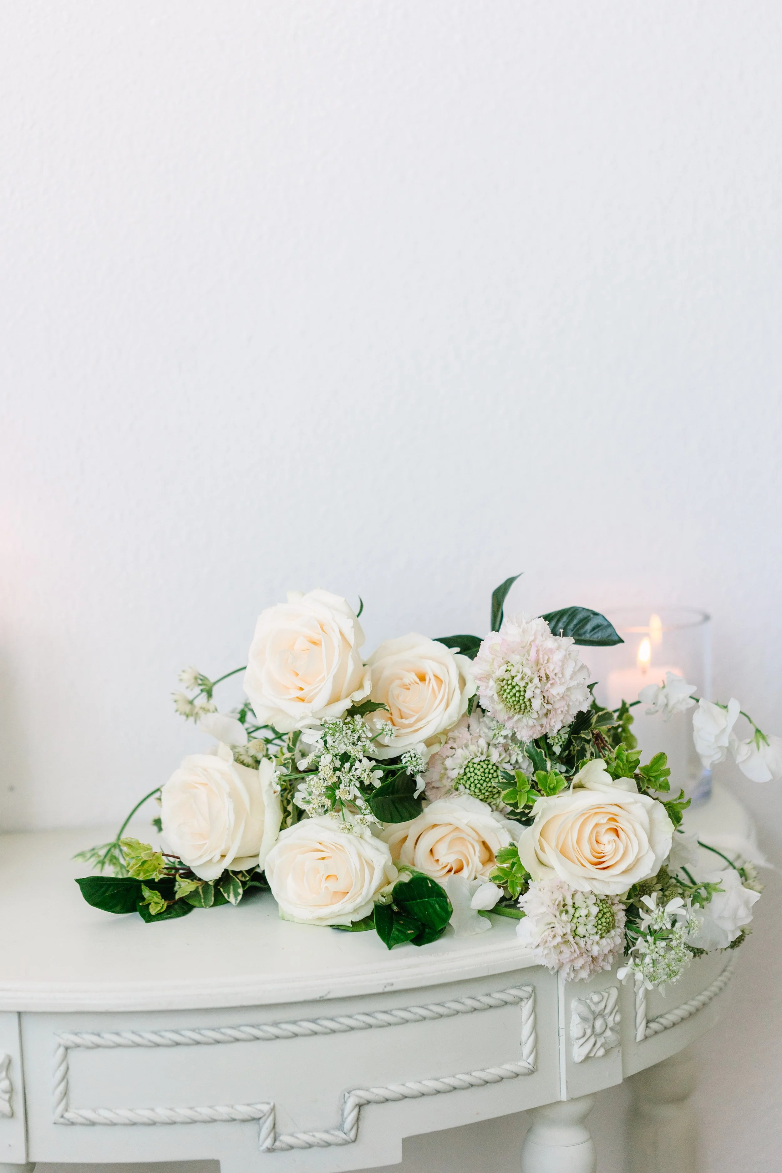 A bouquet of white roses, pink flowers, and greenery on a white vintage table, with a lit candle in the background.