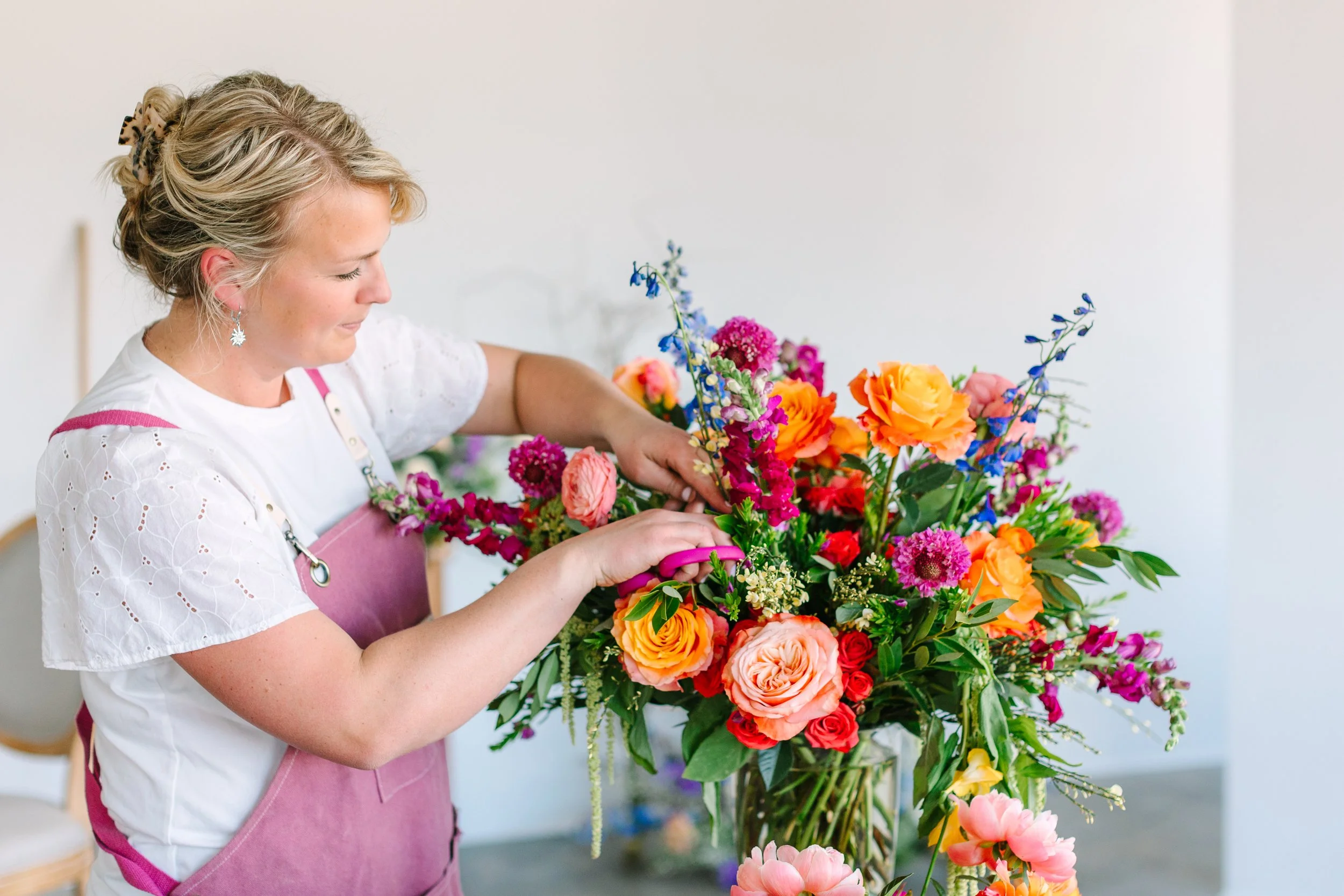 A woman in a pink apron arranging a colorful bouquet of flowers, including roses, snapdragons, and other blooms, in a bright, minimalistic room.