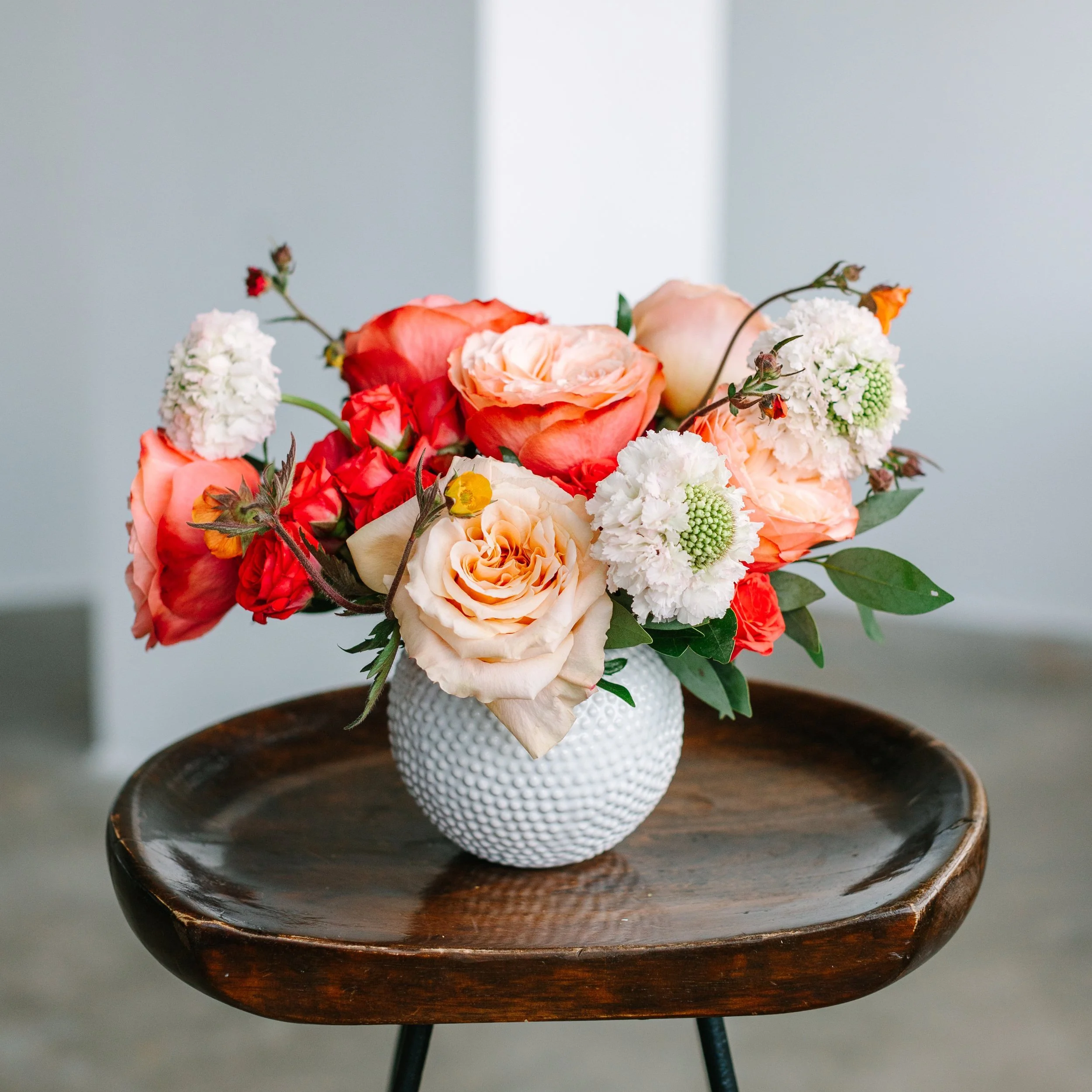 A bouquet of various flowers, including roses, ranunculus, and other blooms, arranged in a textured white vase on a wooden side table.