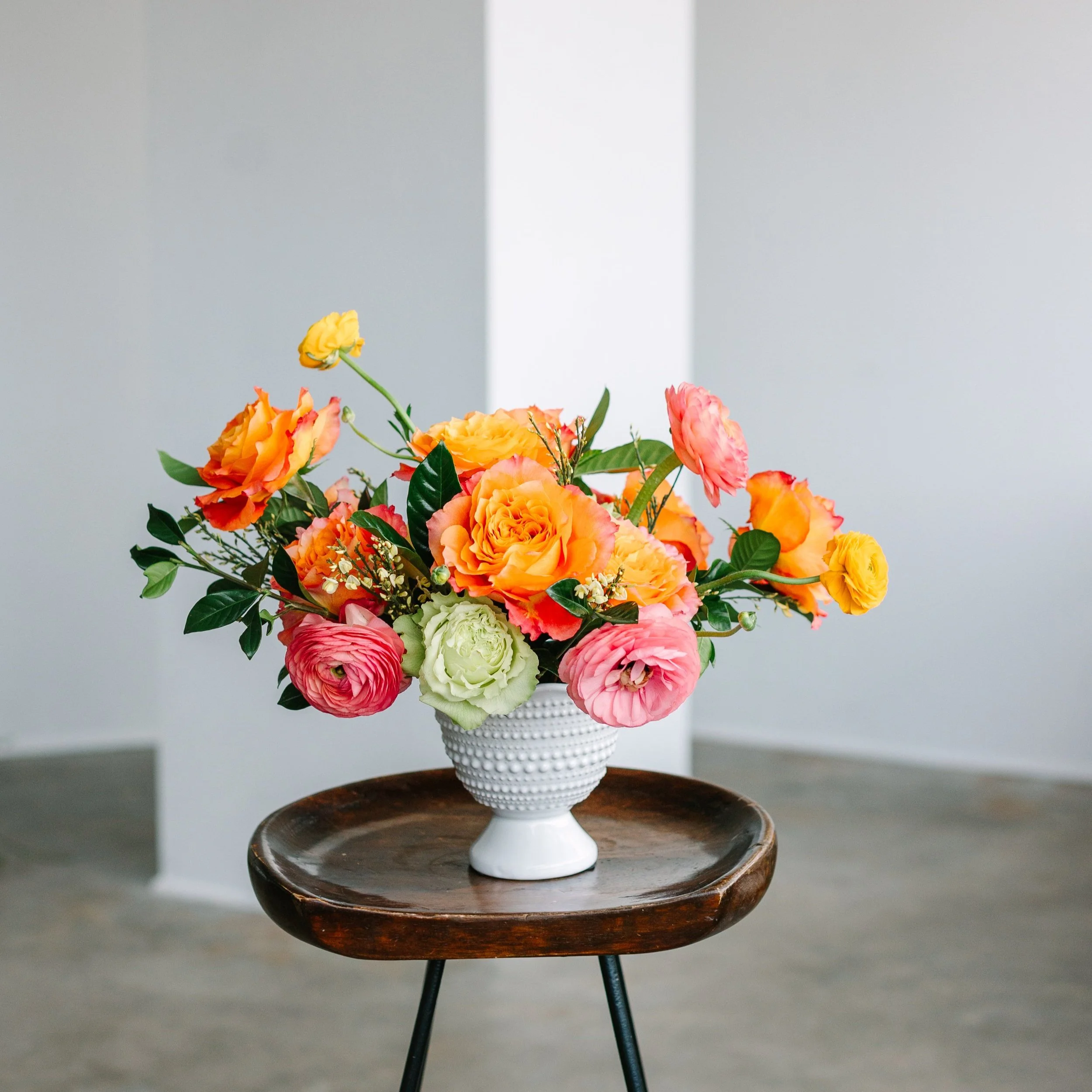 A colorful bouquet of orange, pink, yellow, and white flowers in a white ceramic vase on a dark wooden round table.