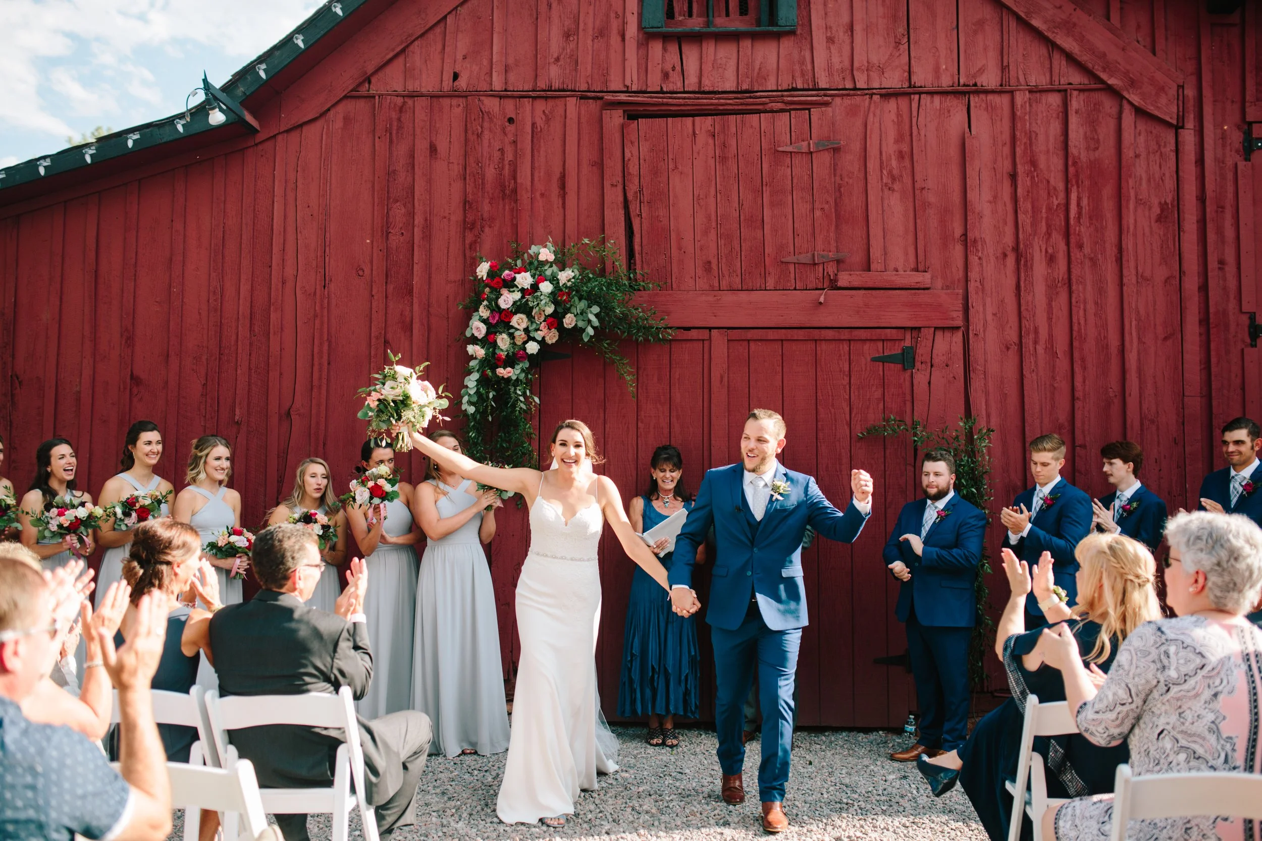 A wedding ceremony outdoors with a group of bridesmaids and groomsmen, the bride in a white dress, and the groom in a blue suit, holding hands and celebrating in front of a red barn with floral decorations.