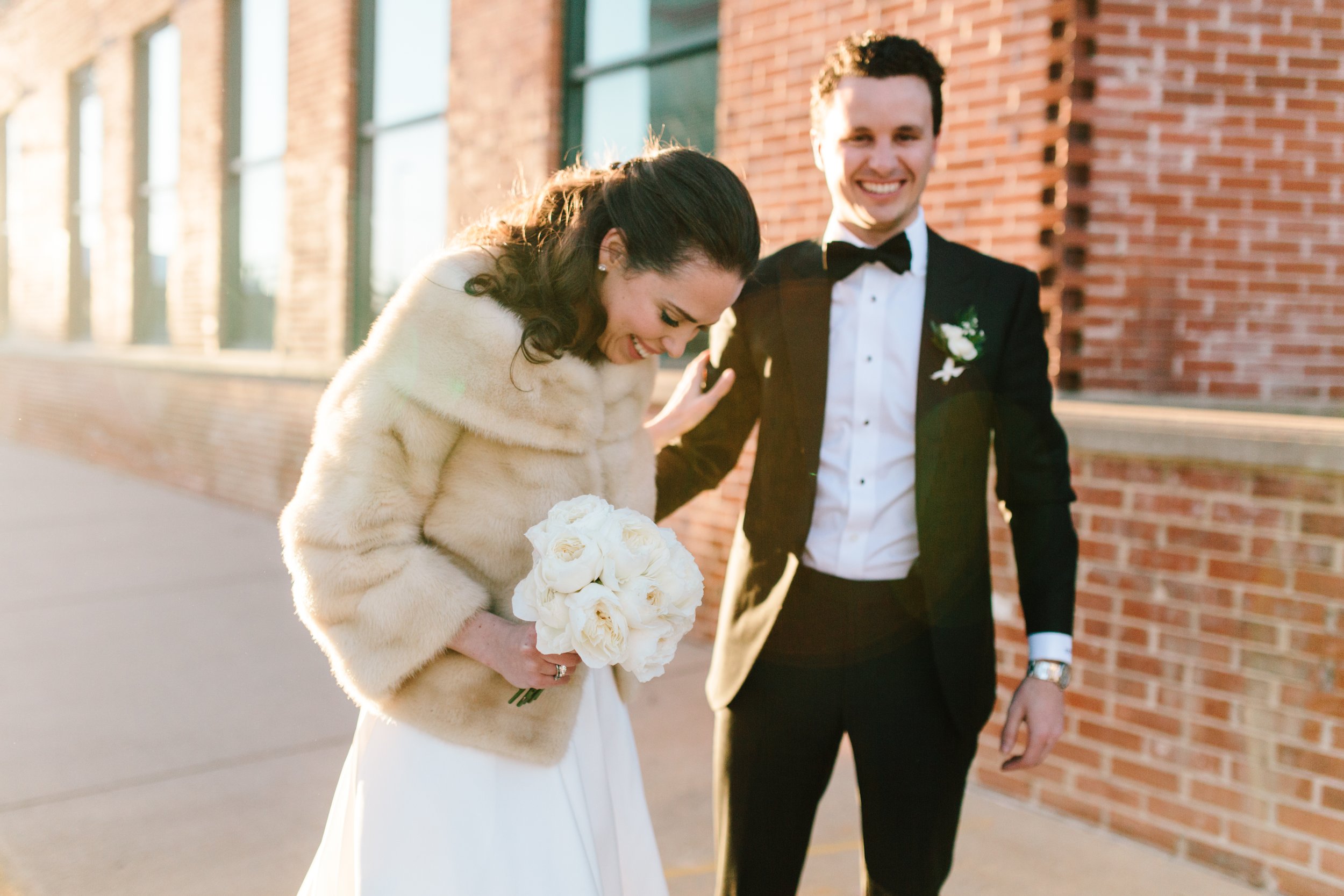 A bride dressed in a white gown and fur coat holding a bouquet of white roses, and a groom in a black tuxedo with a white shirt and black bow tie, standing outdoors against a brick wall during sunset, smiling and sharing a joyful moment.