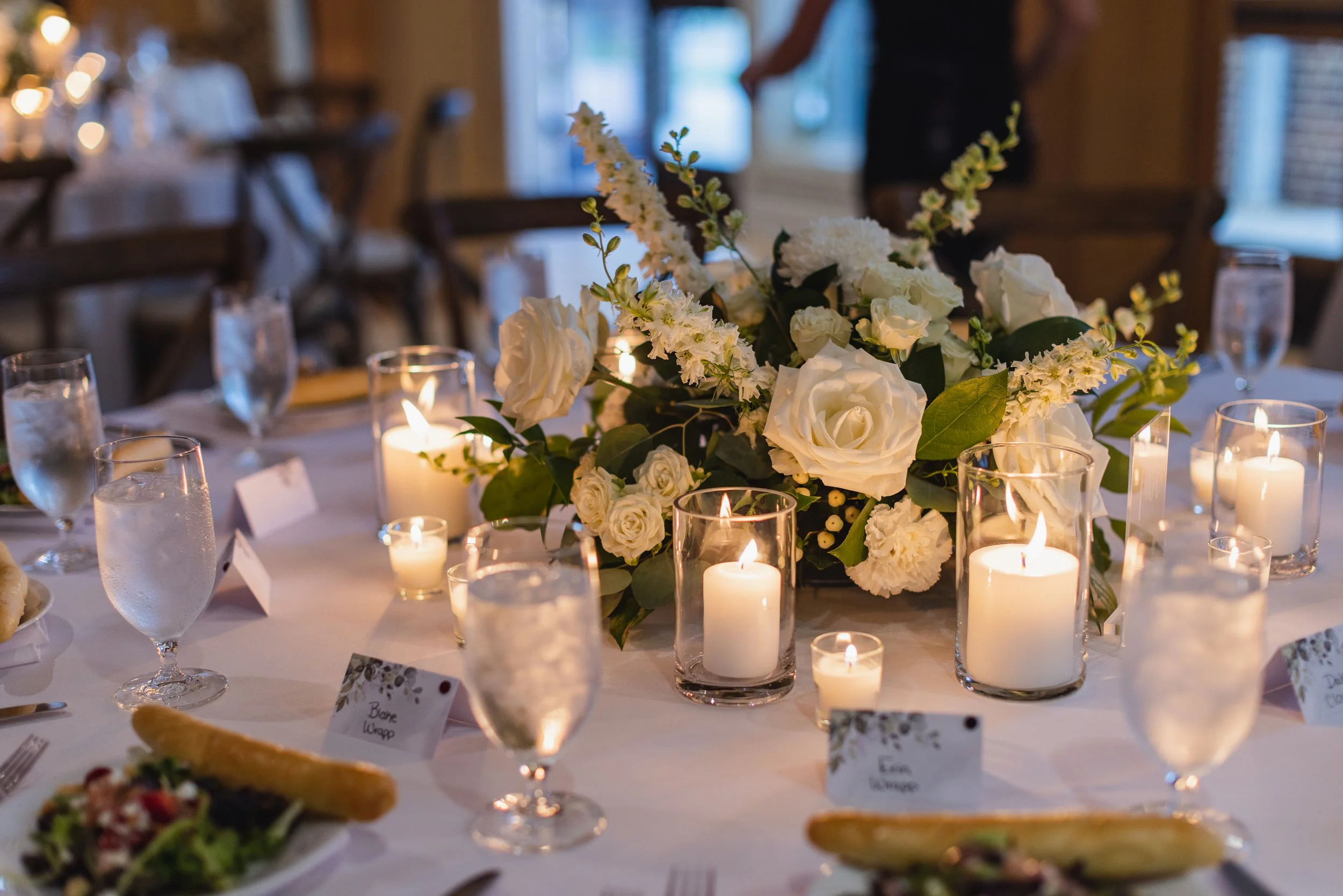 A wedding reception table decorated with a large centerpiece of white roses, hydrangeas, and greenery, surrounded by lit candles in glass holders and water glasses with ice.