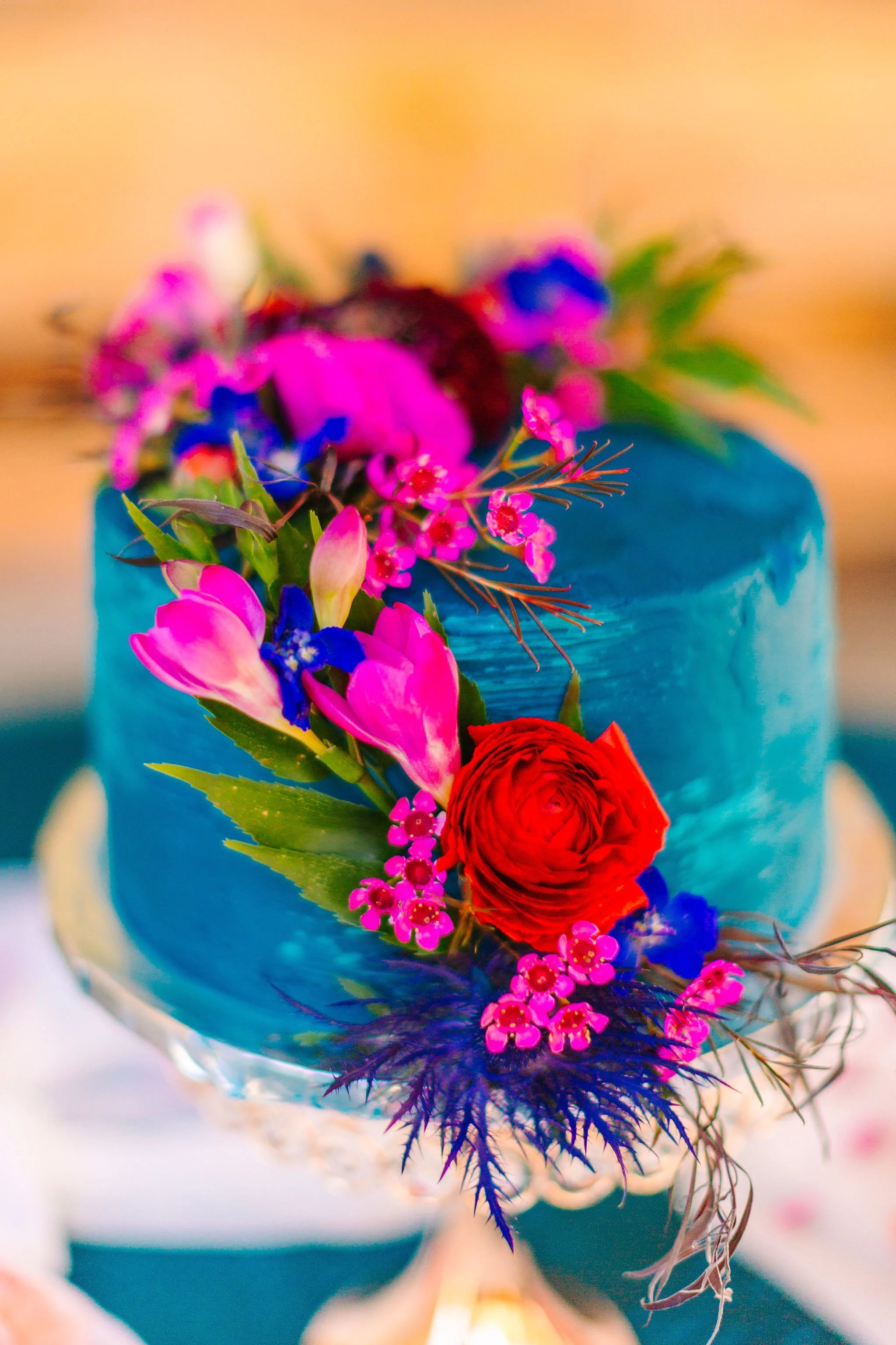 A vibrant cake decorated with pink, blue, and red flowers on top and cascading down the side.