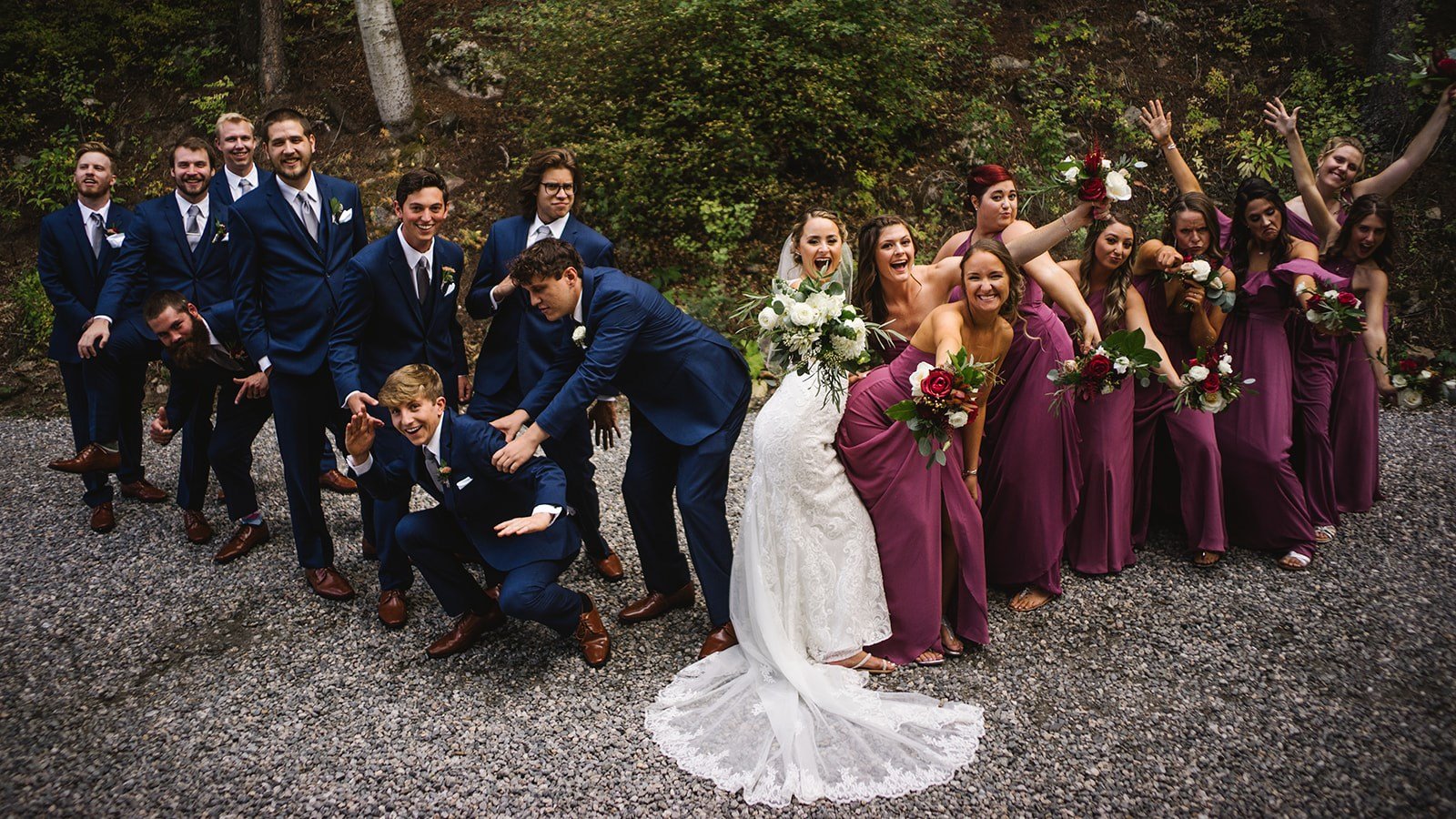 Group of wedding party members, including bride, groom, and bridesmaids in purple dresses, posing outdoors on a gravel path with a wooded background, smiling and making playful gestures.