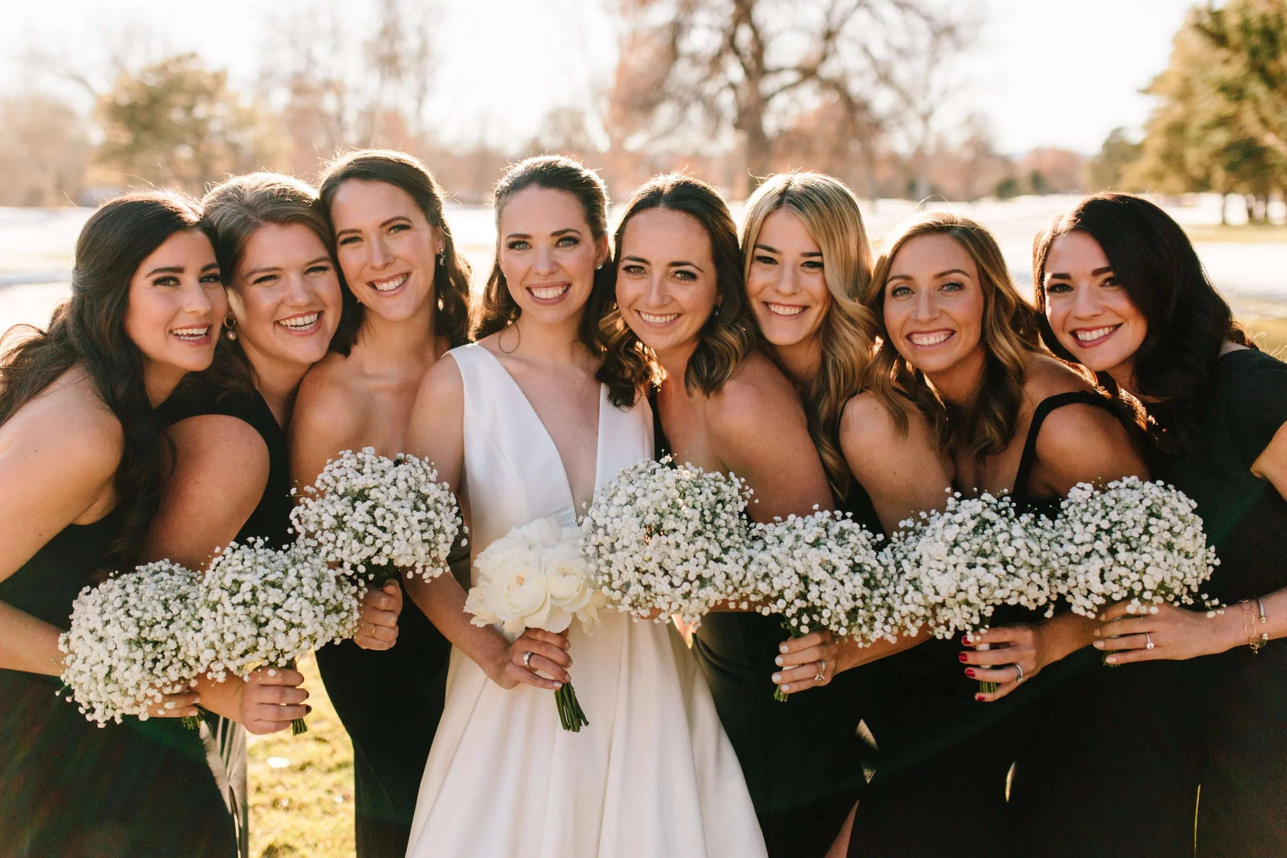 Group of women, including a bride in a white dress, smiling and holding bouquets of white flowers outdoors during sunset with trees in the background.