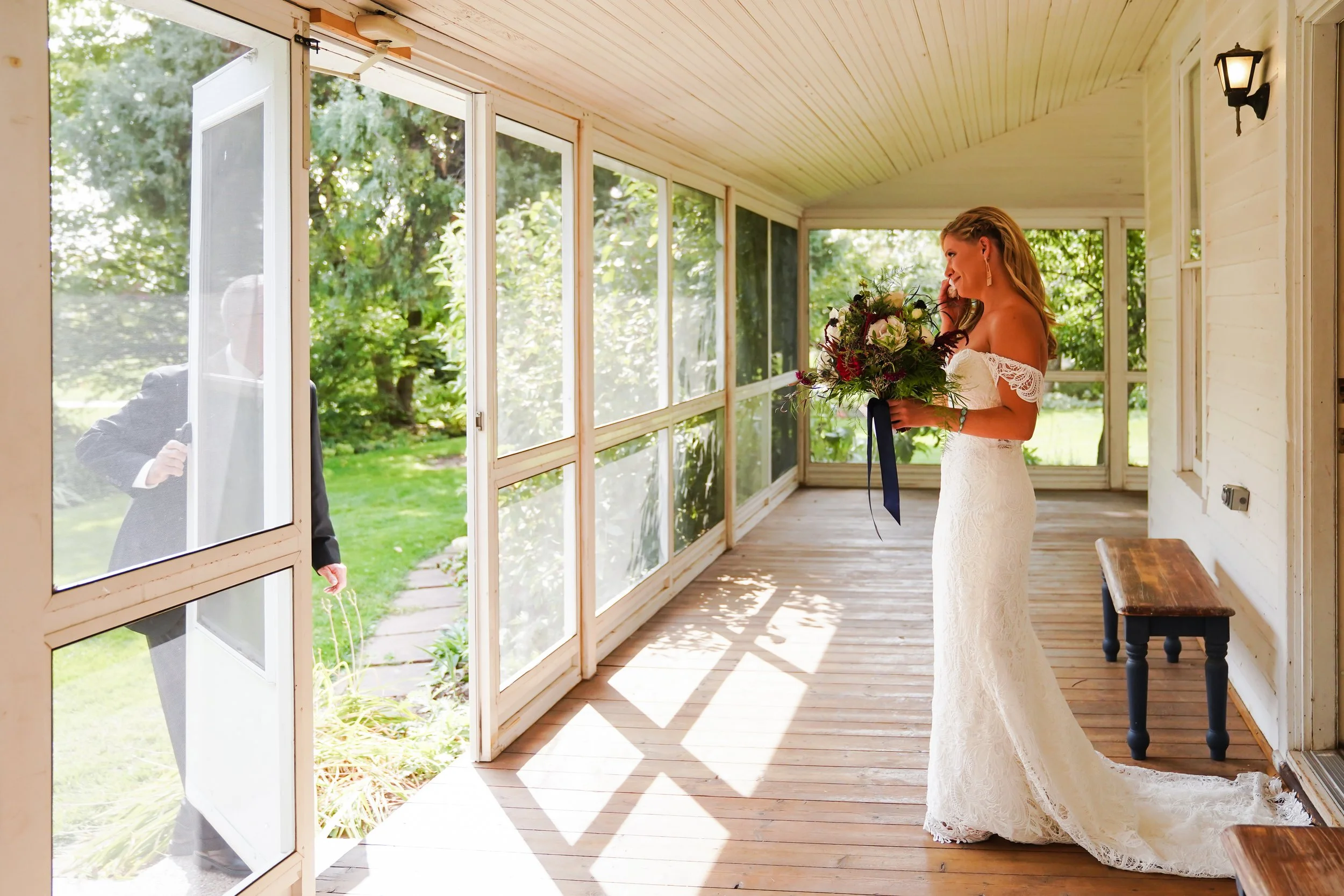 A bride in a white lace wedding dress holding a bouquet, standing on a sunlit porch, looking at a man outside through a screen door.