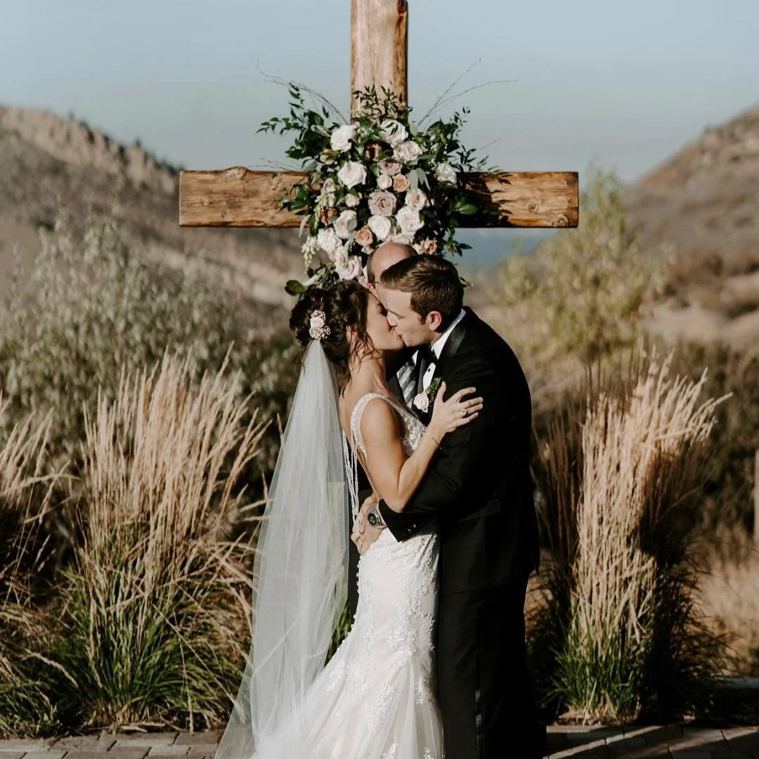 A bride and groom sharing a kiss at their outdoor wedding ceremony, with a wooden cross decorated with flowers behind them and natural landscape in the background.