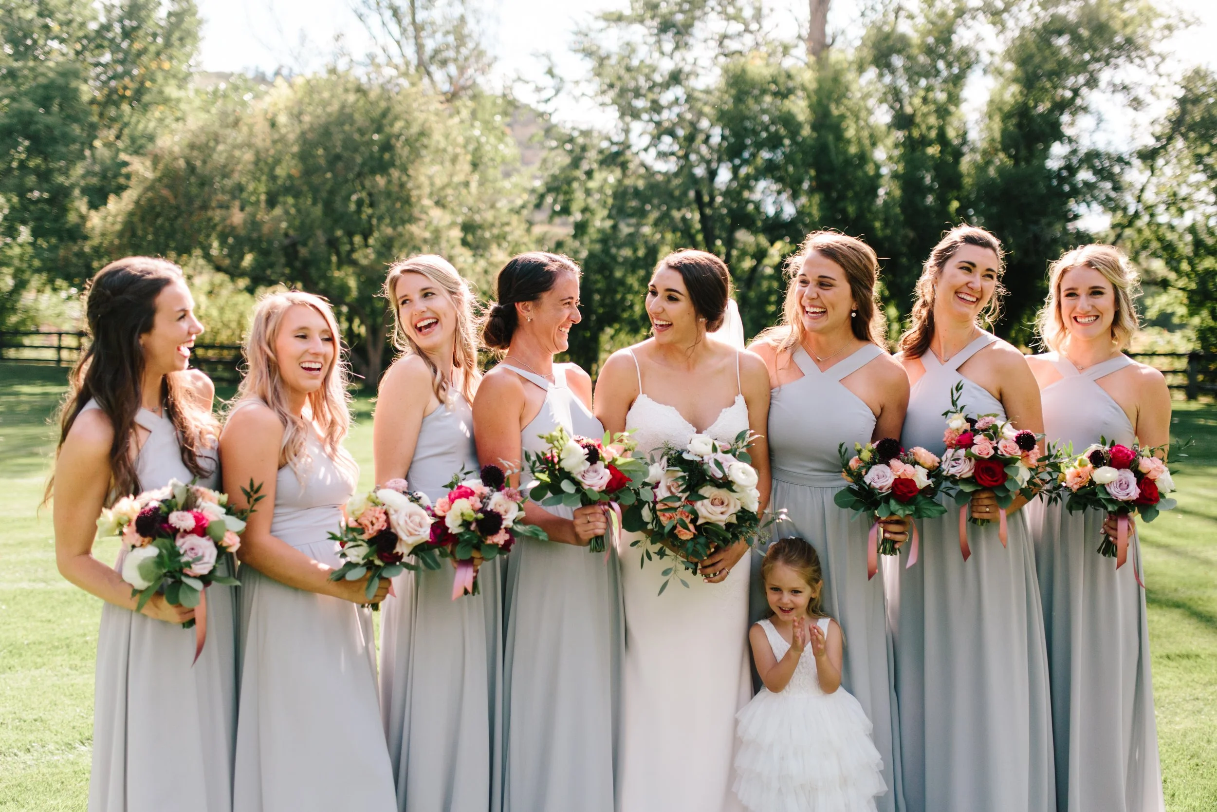 A bride and bridesmaids standing outdoors, each holding a bouquet of flowers, smiling and looking at each other during a wedding celebration.