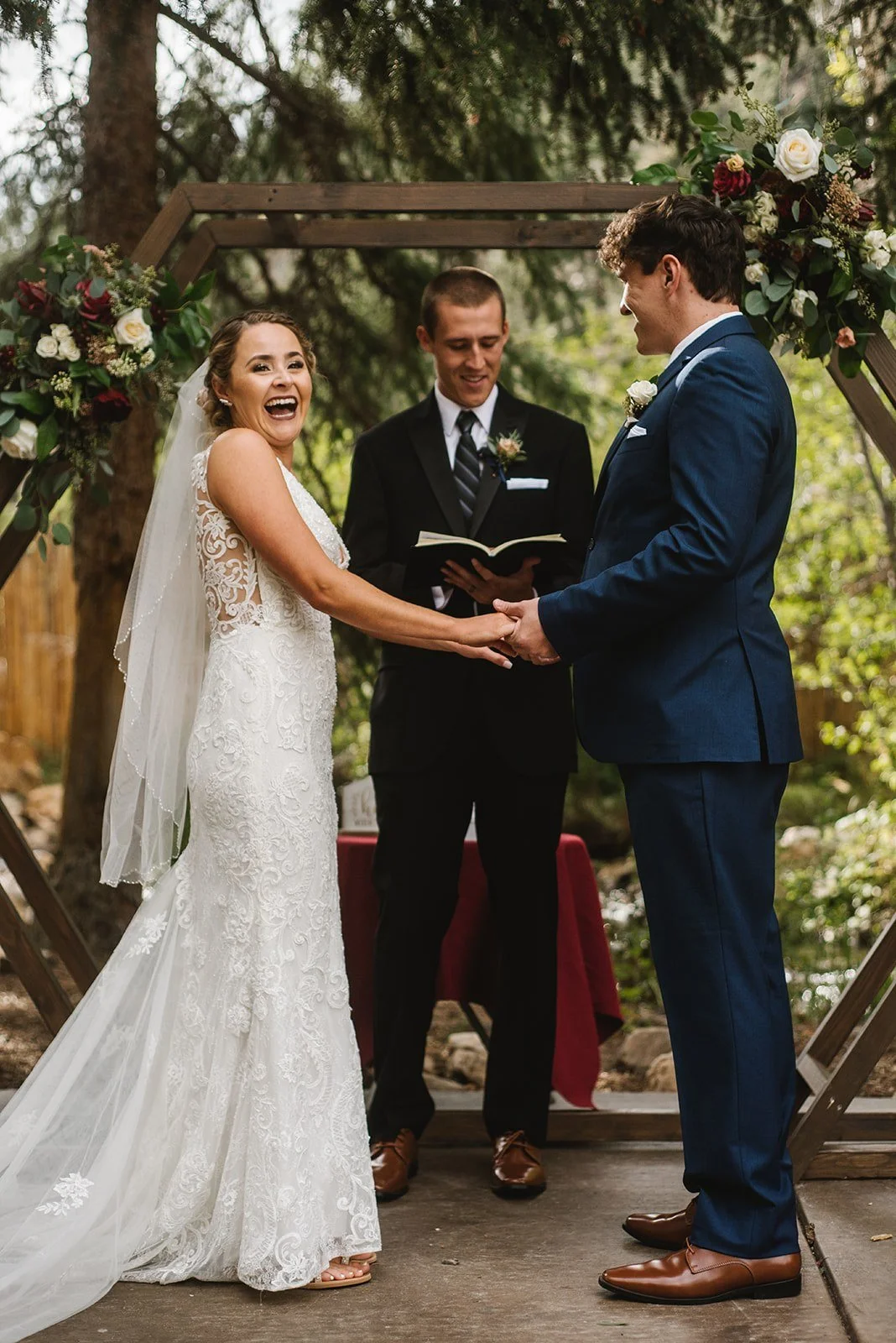 Bride and groom holding hands during their outdoor wedding ceremony, with officiant reading from a book, under a wooden arch decorated with flowers, in a lush green forest setting.