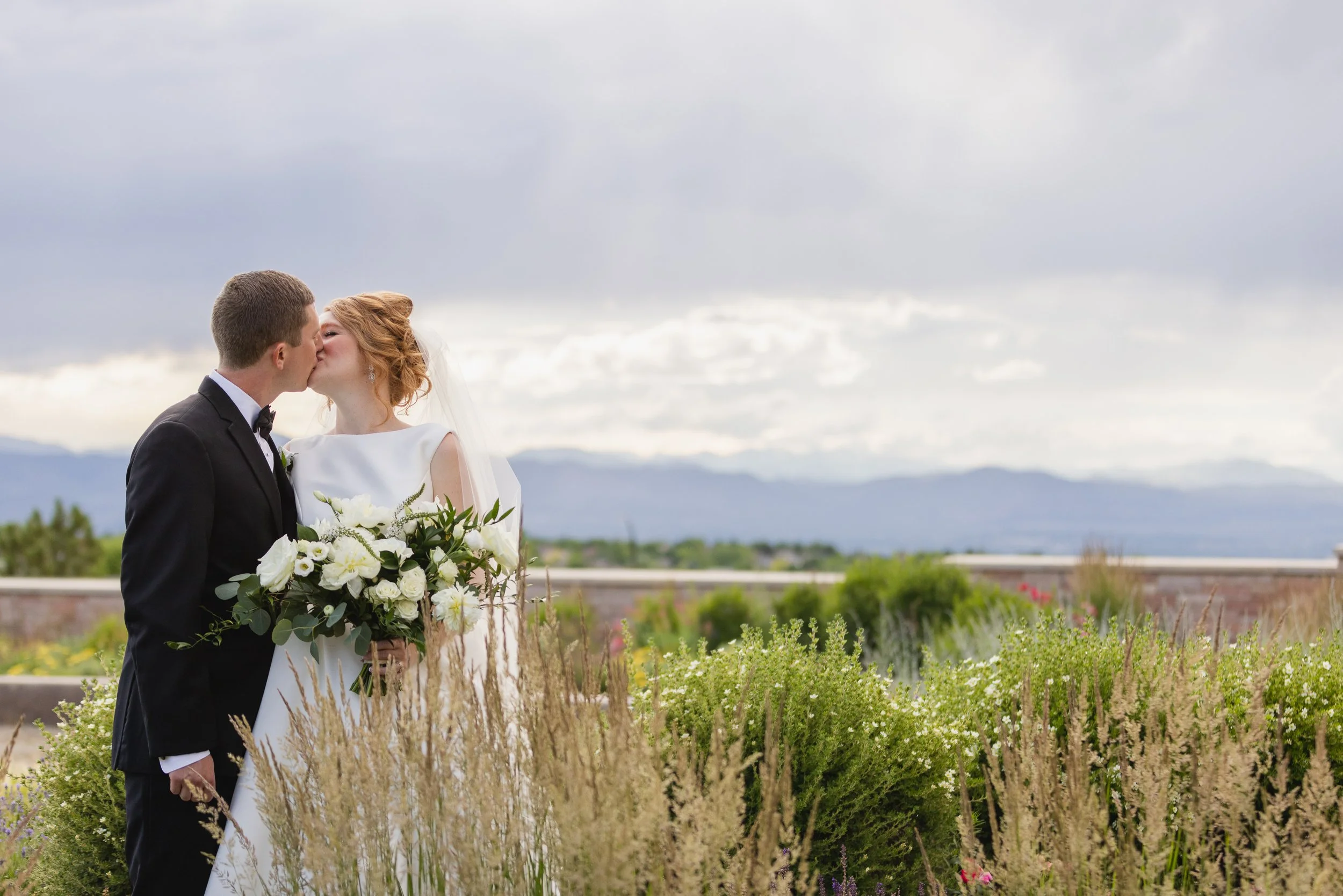 A bride and groom sharing a kiss outdoors on their wedding day, with the bride holding a bouquet of white flowers, greenery, and standing in front of tall grass and flowering bushes, with mountains and cloudy sky in the background.