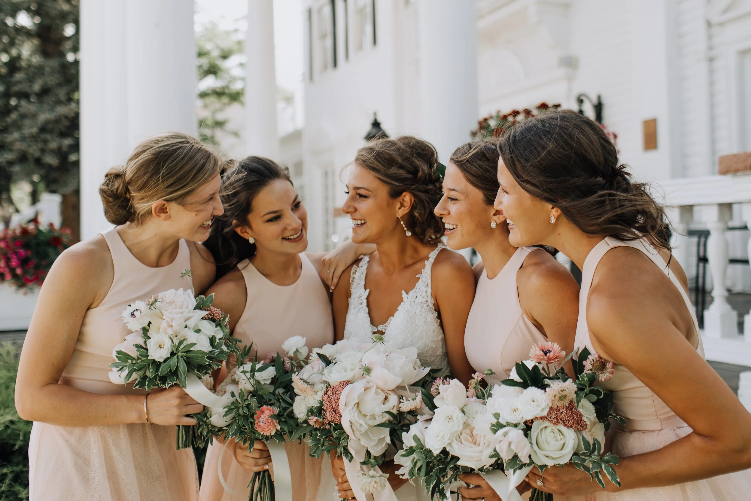 Bride and five bridesmaids in light pink dresses smiling and holding bouquets outside a white house