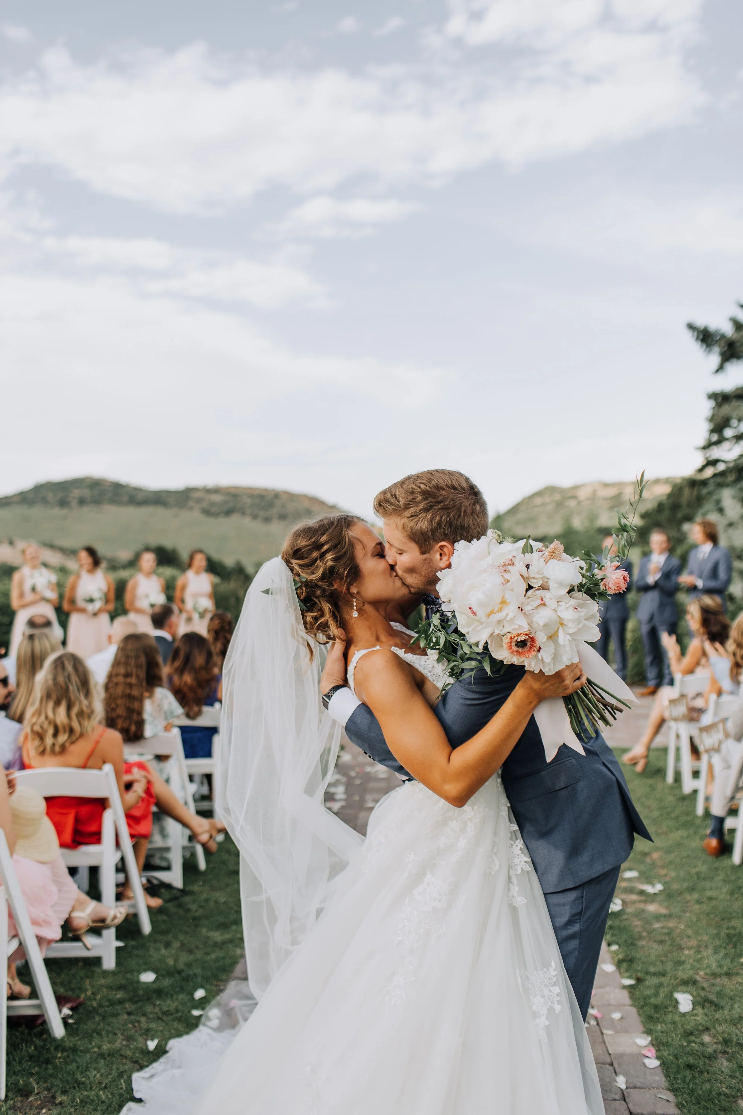 A newly married couple sharing a kiss at their wedding ceremony outdoors, surrounded by guests and scenic hills in the background.