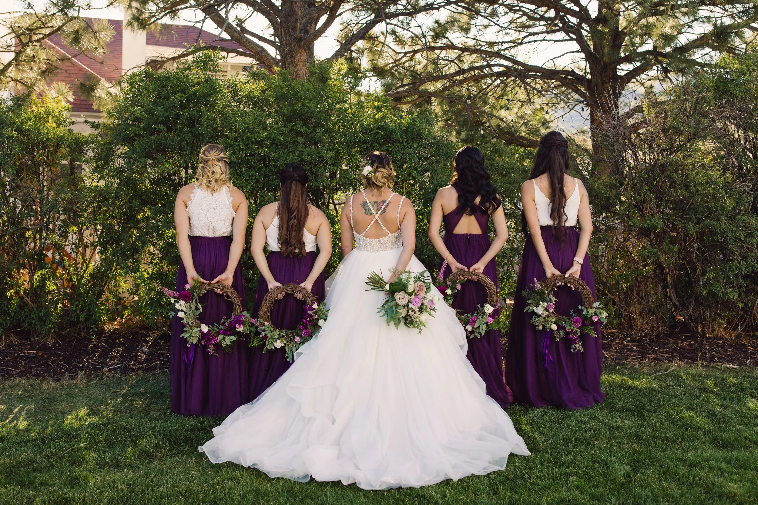 Bride and five bridesmaids standing outdoors with their backs, facing a green hedge and trees, on a grassy area during a wedding.