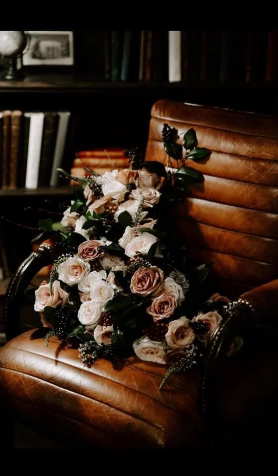 A bouquet of pink and white roses with dark greenery resting on a brown leather chair in front of a dark bookshelf.