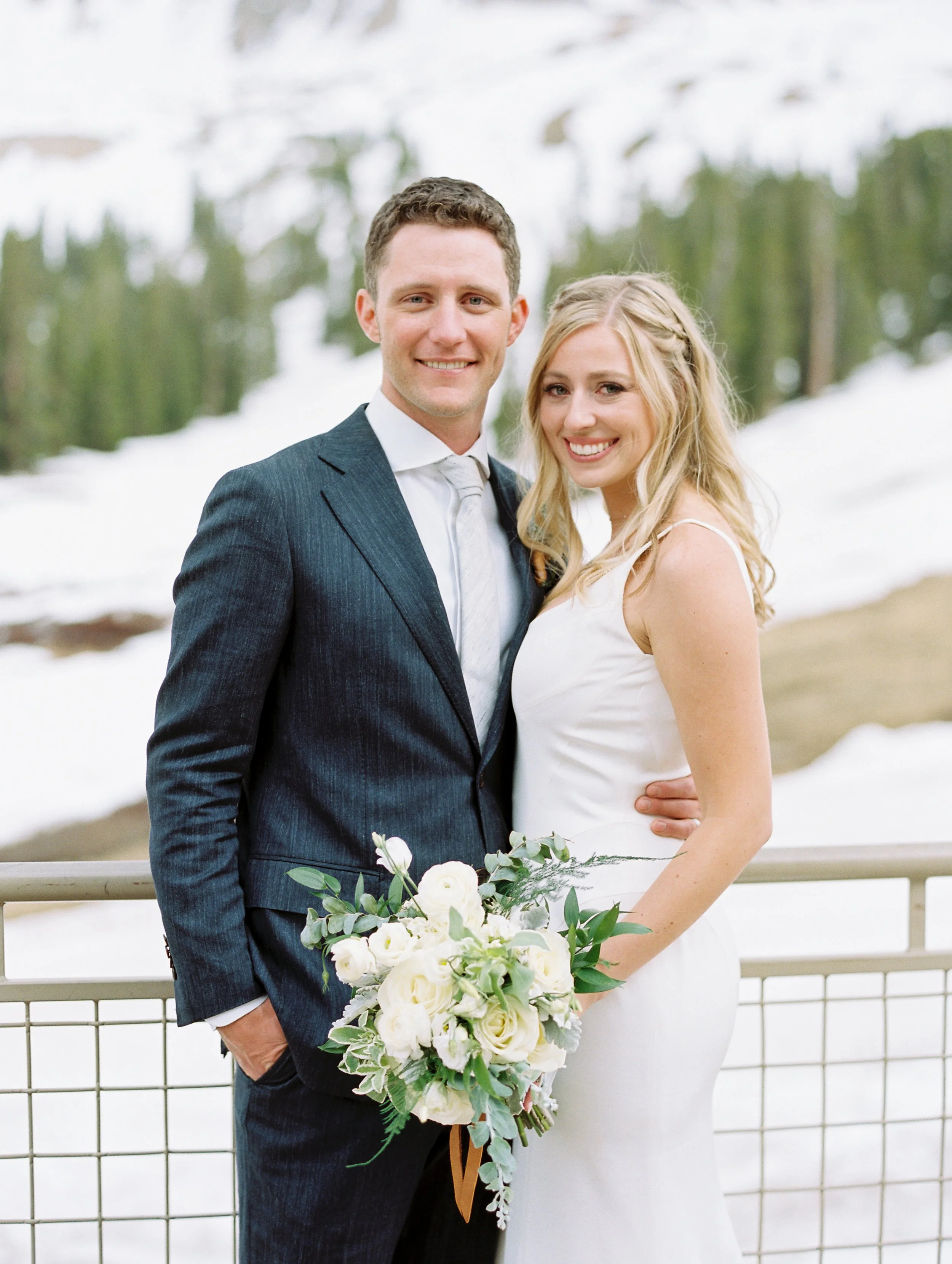 A newlywed couple standing outdoors with snow-covered mountains and trees in the background. The groom wears a dark suit with a white shirt and tie, and the bride wears a white dress holding a bouquet of white flowers and greenery.