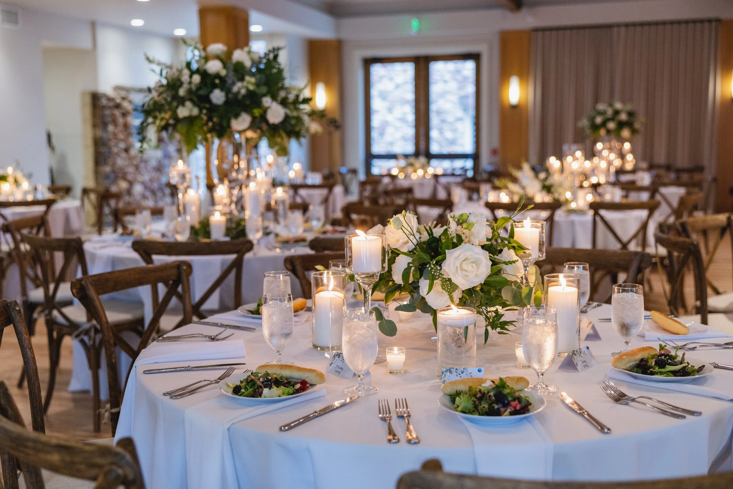 Elegant banquet hall decorated for a wedding reception with round tables covered in white tablecloths, floral centerpieces, candles, and place settings of plates, utensils, glasses and bread. Comfortable wooden chairs surround each table.