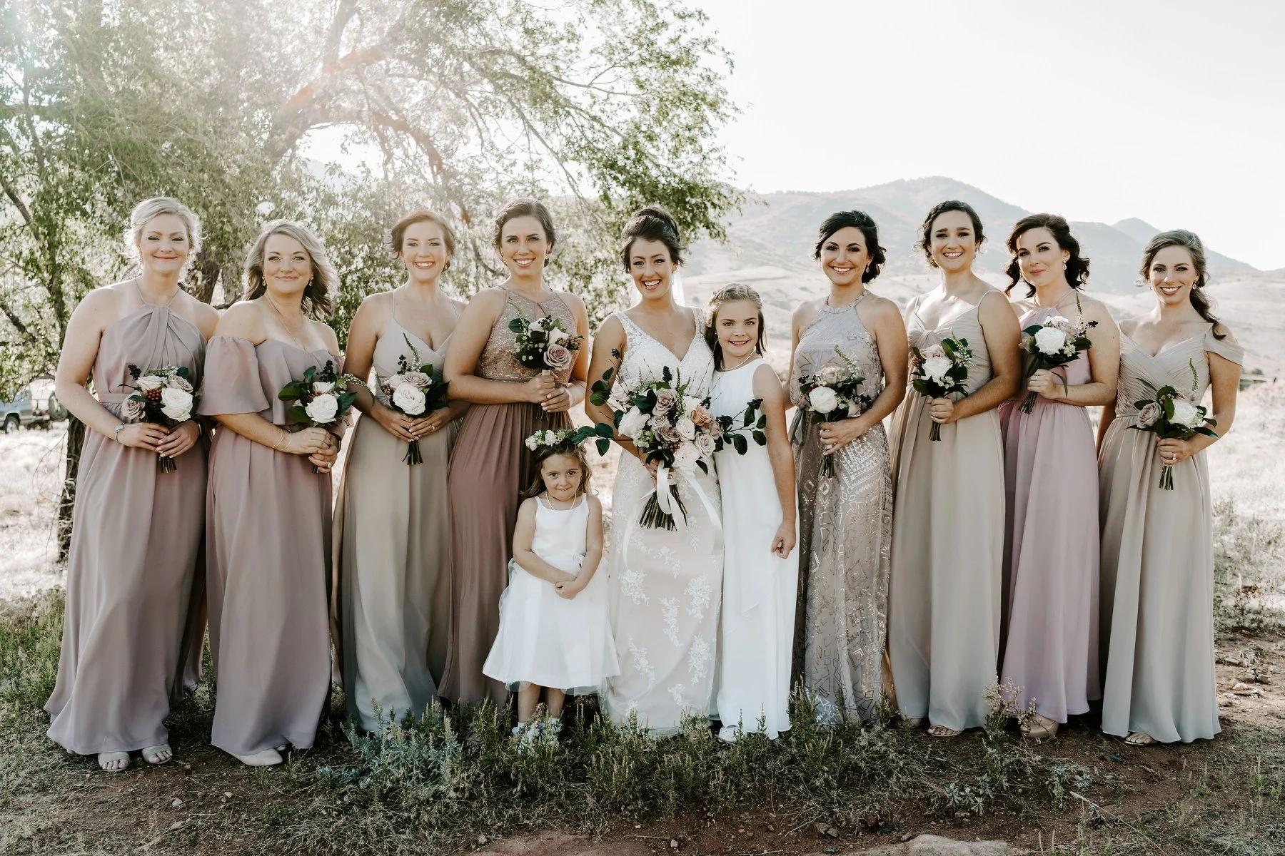 Group of women and young girls in formal dresses, holding bouquets, outdoors in natural setting with mountains in the background.