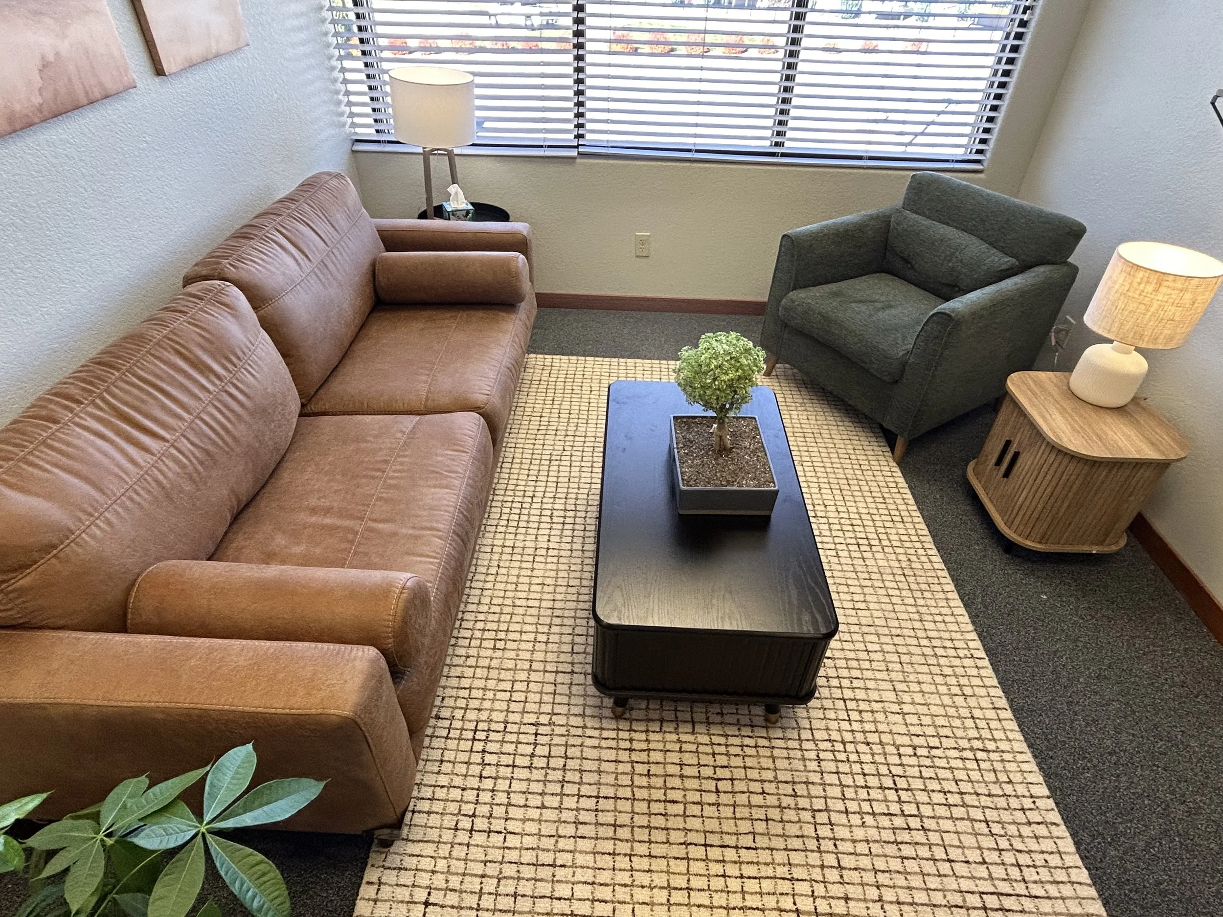Living room with brown leather sofa, green armchair, black coffee table with a potted plant, and wooden side table with a lamp, window with blinds.