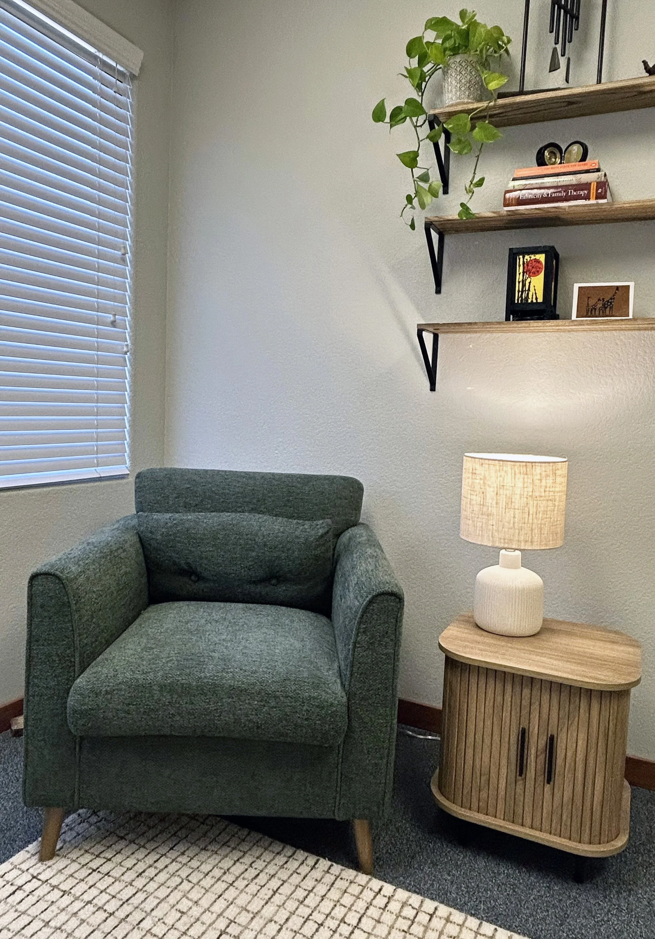 A cozy corner with a green upholstered armchair, a wooden side table with a white ceramic lamp, and wall-mounted wooden shelves holding books, small decorative items, and a potted plant. There is a window with blinds to the left.