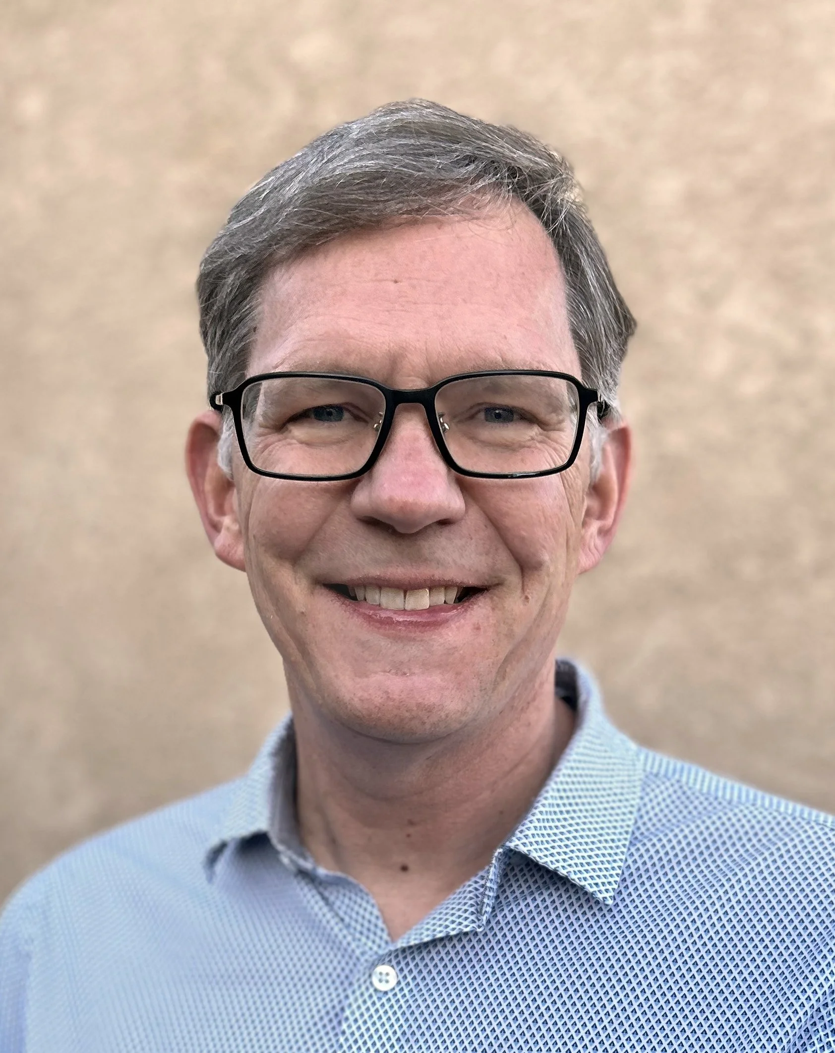 A smiling man wearing glasses and a light blue collared shirt, against a beige textured background.