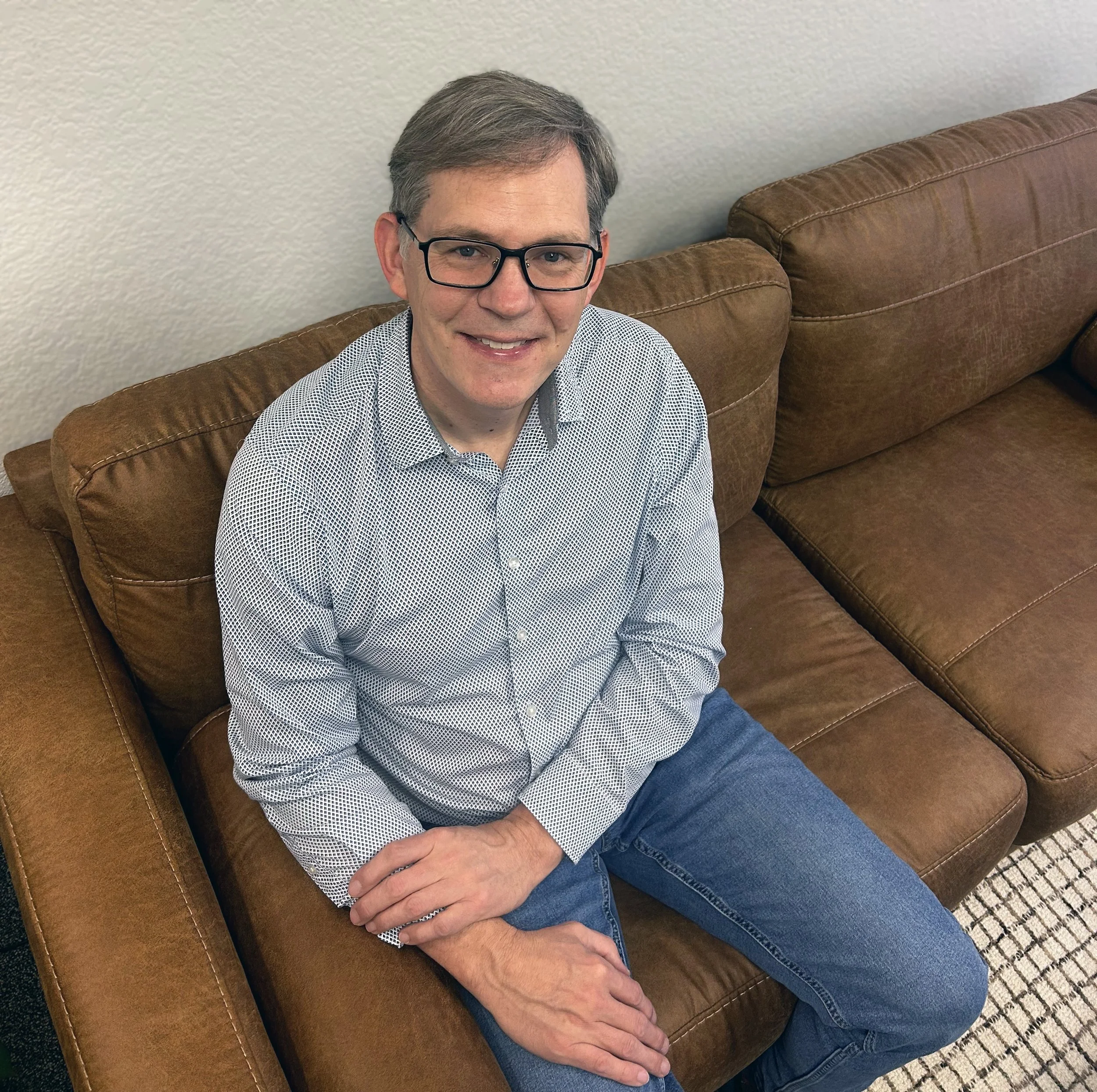 A man with glasses wearing a patterned button-up shirt and jeans sitting on a brown leather couch, smiling, in a room with a light-colored wall.