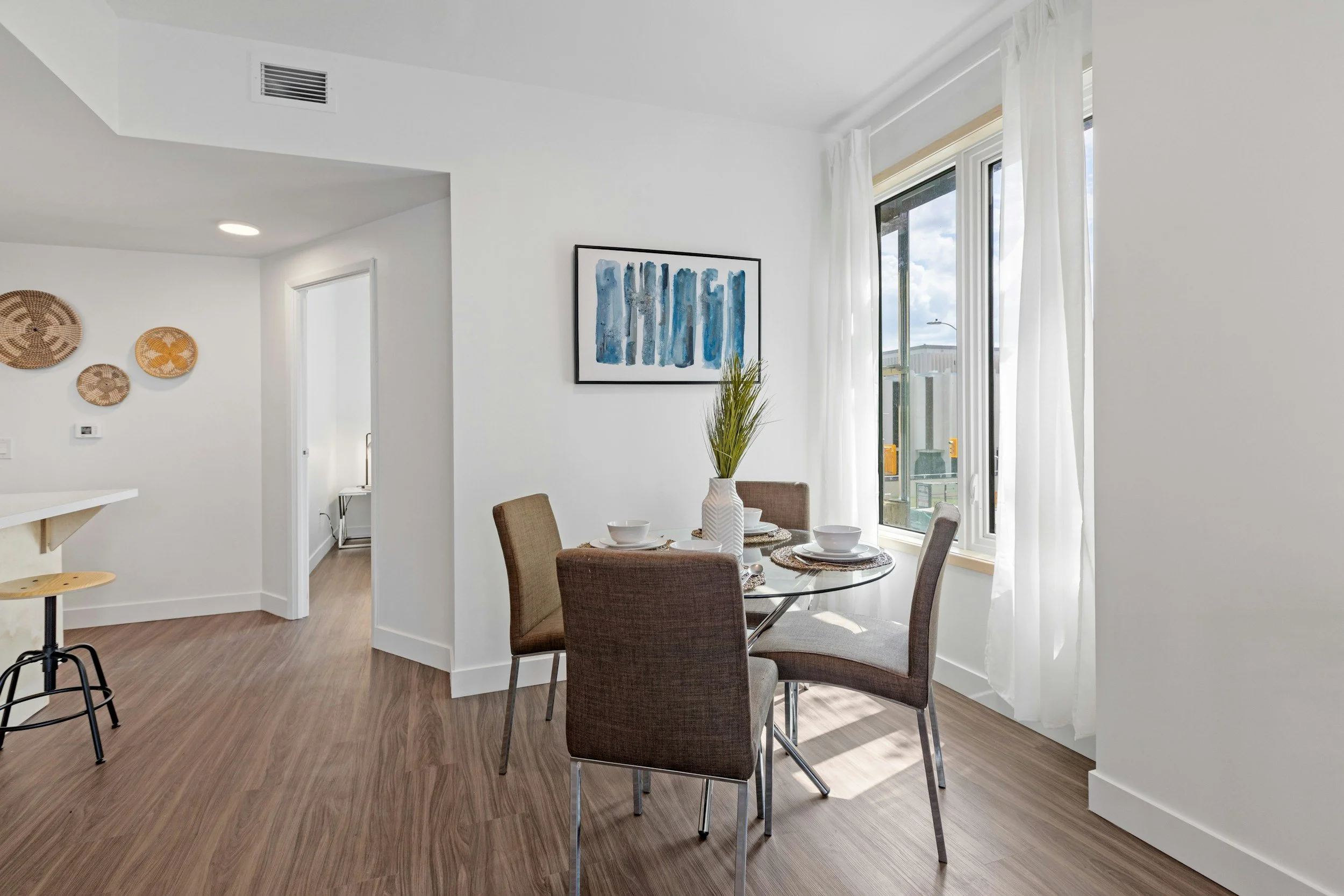 Dining area with a round glass table, four brown upholstered chairs, white curtains, a window letting in natural light, and a white vase with green plants on the table.